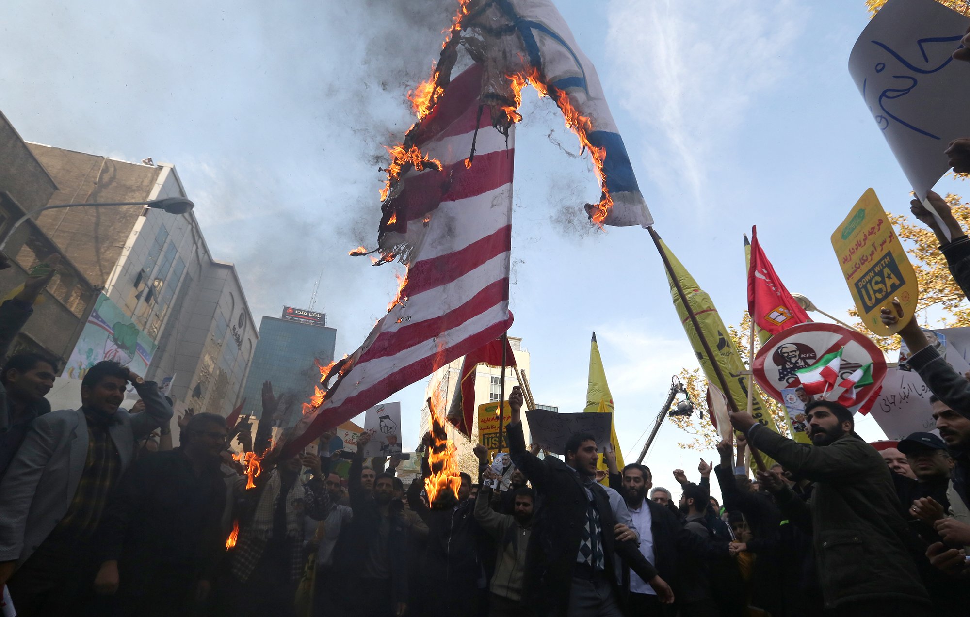 L'image montre une scène de manifestation où des personnes sont rassemblées pour protester. Au centre, un drapeau semble être en feu, ce qui attire l'attention des manifestants. Ils tiennent également des pancartes avec différents slogans et symboles. L'atmosphère est tendue, et on ressent un fort sentiment d'engagement parmi les participants. L'arrière-plan présente des bâtiments urbains, suggérant une grande ville.