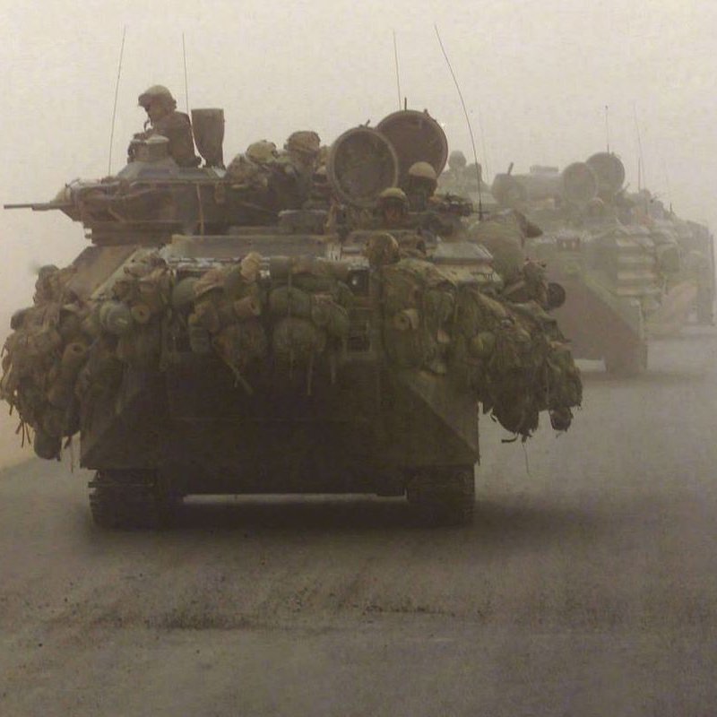 L'image montre des véhicules militaires, probablement des chars, se déplaçant dans un environnement poussiéreux et brumeux. On peut voir des soldats à bord des chars, et ces derniers semblent être chargés avec des équipements. L'atmosphère est chargée, créant une impression de tension et d'intensité, probablement en lien avec une situation de conflit ou d'opération militaire.