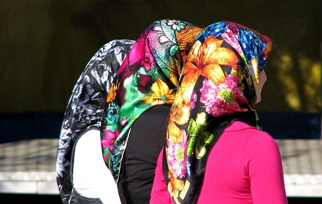 L'image montre le dos de trois femmes portant des foulards colorés. Chaque foulard est orné de motifs fleuris et vibrants, offrant un contraste frappant avec leurs vêtements. Les cheveux des femmes sont couverts, laissant entrevoir seulement leurs silhouettes. L'ambiance semble vivante et pleine de couleurs, soulignant la richesse culturelle des accessoires.