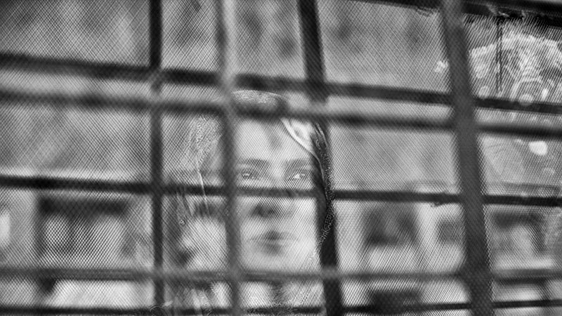 L'image montre une femme qui apparaît derrière une grille. Le contraste entre le flou et les barreaux de la grille donne une impression de séparation ou d'enfermement. La femme semble contemplative, et son expression peut évoquer des émotions profondes. Les nuances de noir et blanc ajoutent une atmosphère mélancolique à la scène.