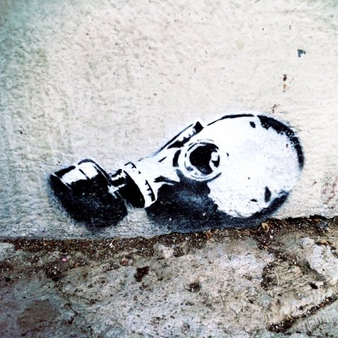 L'image montre un graffiti représentant une tête de mort portant un masque à gaz. Ce dessin est peint en noir et blanc et est appliqué sur un mur, près du sol. Le contraste entre le noir et le blanc met en évidence les contours du masque et de la tête, créant une image saisissante et inquiétante. Le style est à la fois moderne et urbain, typique des œuvres de street art.