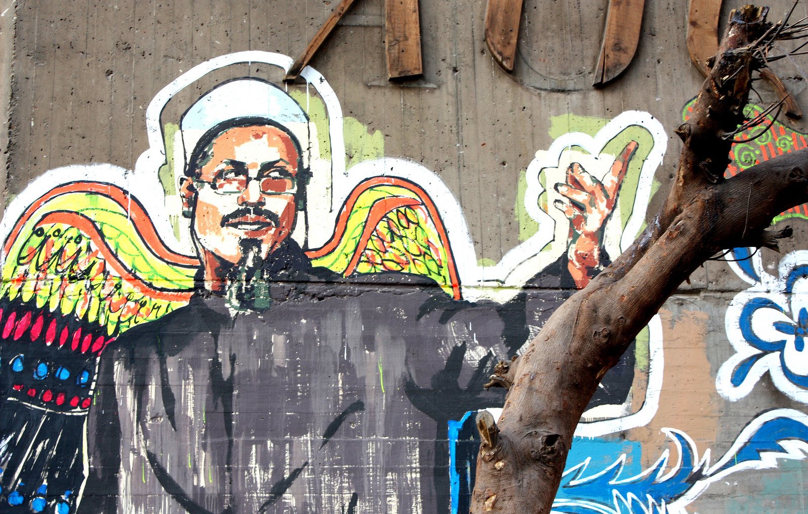 L'image représente une fresque murale colorée. On y voit un homme aux épaules larges, vêtu d'une tunique noire et d'un chapeau traditionnel. Il a un regard expressif et semble faire un geste de la main, comme s'il s'adressait à quelqu'un. En arrière-plan, des motifs vibrants et contrastés, dont des ailes colorées, ajoutent une dimension artistique à la composition. Un arbre, avec des branches dénudées, est également présent au premier plan, soulignant l'environnement urbain de la fresque.