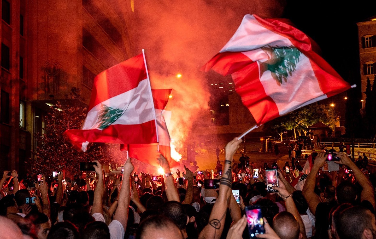 تظهر الصورة تجمعًا حاشدًا في أحد الشوارع، حيث يحمل المتظاهرون أعلام لبنان. تضيء الأجواء بألوان النيران والدخان، مما يخلق جوًا احتفاليًا أو تعبيريًا. يُلاحظ أن الكثير من الأشخاص يستخدمون هواتفهم لتسجيل اللحظة أو التقاط الصور، بالإضافة إلى الأضواء الساطعة التي تضيف حيوية للمشهد.
