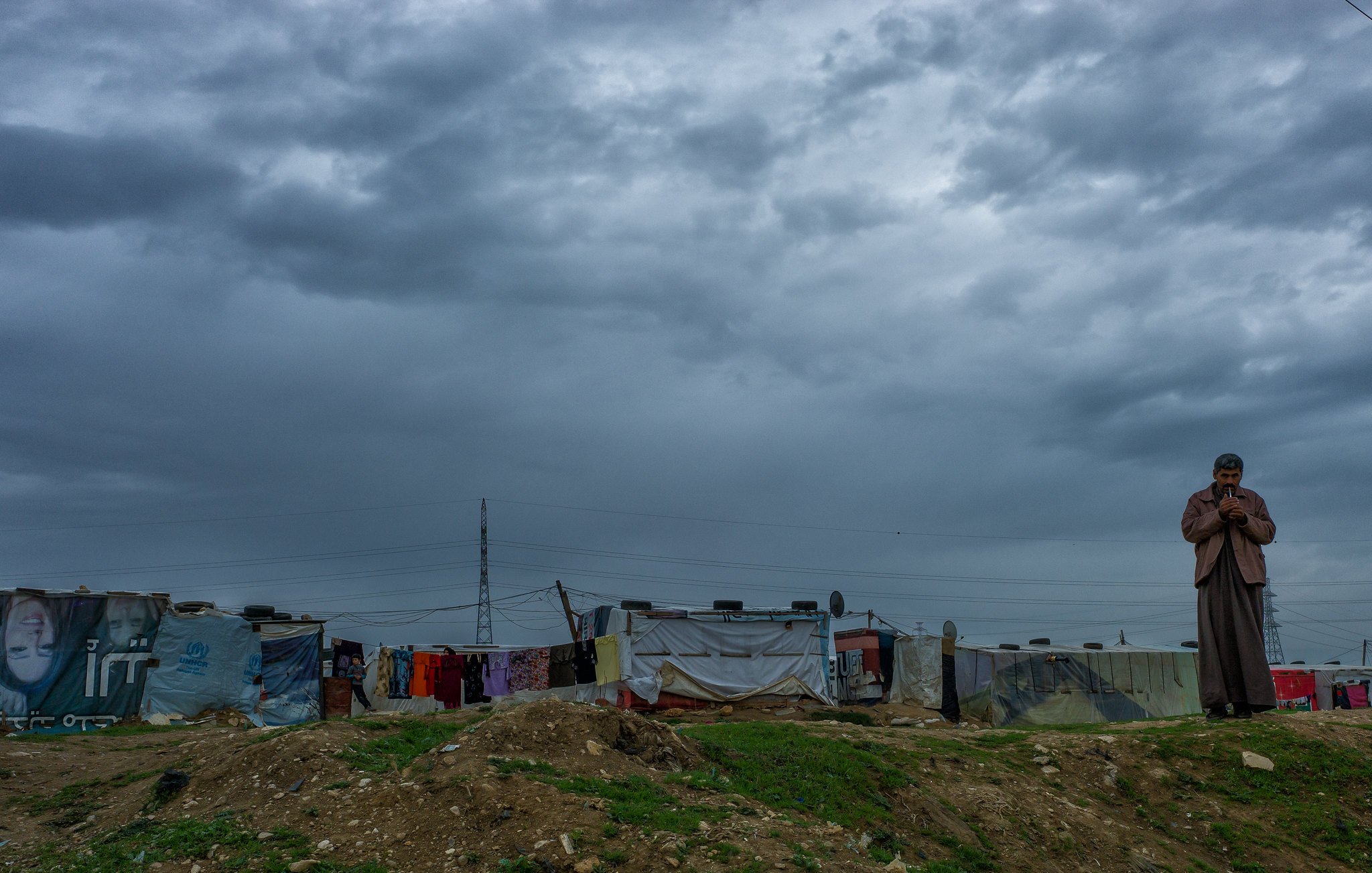 در این تصویر، یک مرد در منطقه‌ای با ساختمان‌های موقتی یا کمپ زندگی می‌کند. آسمان ابری و تیره به نظر می‌رسد و نشان‌دهنده شرایط جوی نامساعد است. در پس‌زمینه، چند چادر یا خانه موقت دیده می‌شود که با پارچه‌ها و وسایل مختلف پوشانده شده‌اند. فضای کلی تصویر حس کمبود و در عین حال استقامت را منتقل می‌کند. مرد به نظر می‌رسد در حال مکالمه یا تفکر است و تصویر کلی فضایی جدی و غم‌انگیز دارد.