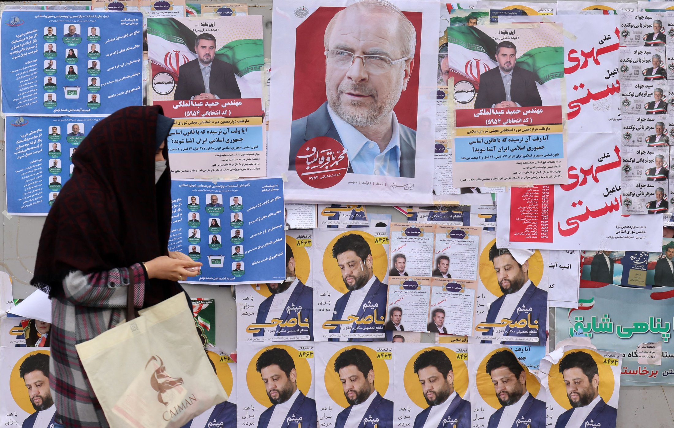 در این تصویر، یک زن که با چادر مشکی پوشیده است، در حال عبور از کنار دیواری است که پر از پوسترهای انتخاباتی و تبلیغاتی است. پوسترها شامل تصاویری از نامزدهای مختلف و شعارهای انتخاباتی هستند. چهره‌ای از یک فرد (احتمالاً نامزد) در مرکز تصویر بسیار مشهود است، در حالی که دیگر پوسترها در اطراف آن به چشم می‌خورند. دیوار پر از رنگ و متن است که به فضای انتخاباتی اشاره می‌کند. زن در حال نگاه کردن به این پوسترها به نظر می‌رسد.