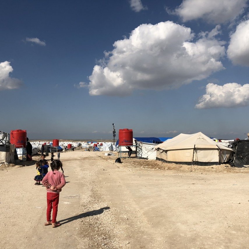 تظهر في الصورة مخيم للاجئين أو النازحين، حيث تتواجد عدة خيام مرتبة على جانبي الطريق. السماء صافية مع وجود سحب بيضاء، والأرض مرصوفة بالحجارة. يمكن رؤية بعض الأطفال يلعبون في المنطقة. يبدو أن المخيم يعاني من ظروف بسيطة، مما يشير إلى حياة موقتة للأشخاص المقيمين فيه.