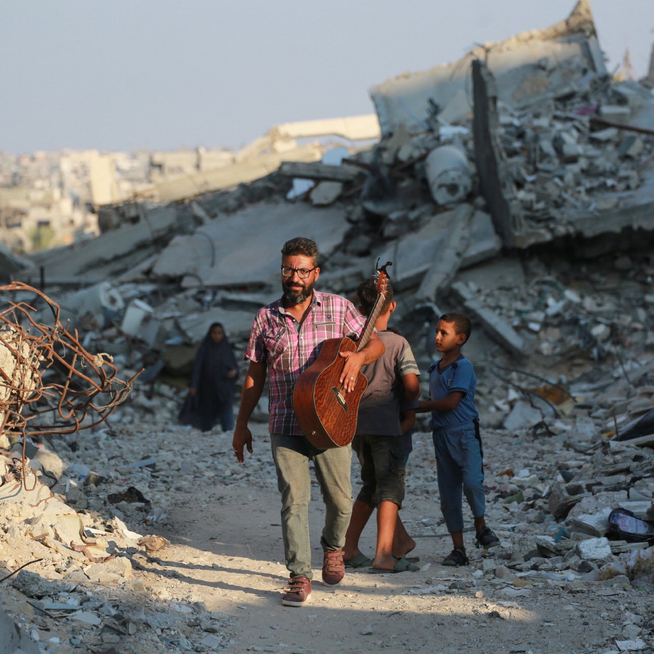 L'image montre un homme marchant à travers des décombres, tenant une guitare. Il est accompagné d'un jeune garçon. L'environnement est en ruines, avec des débris et des structures effondrées autour d'eux. L'atmosphère semble à la fois désolée et résiliente, illustrant un contraste entre la musique et la destruction.
