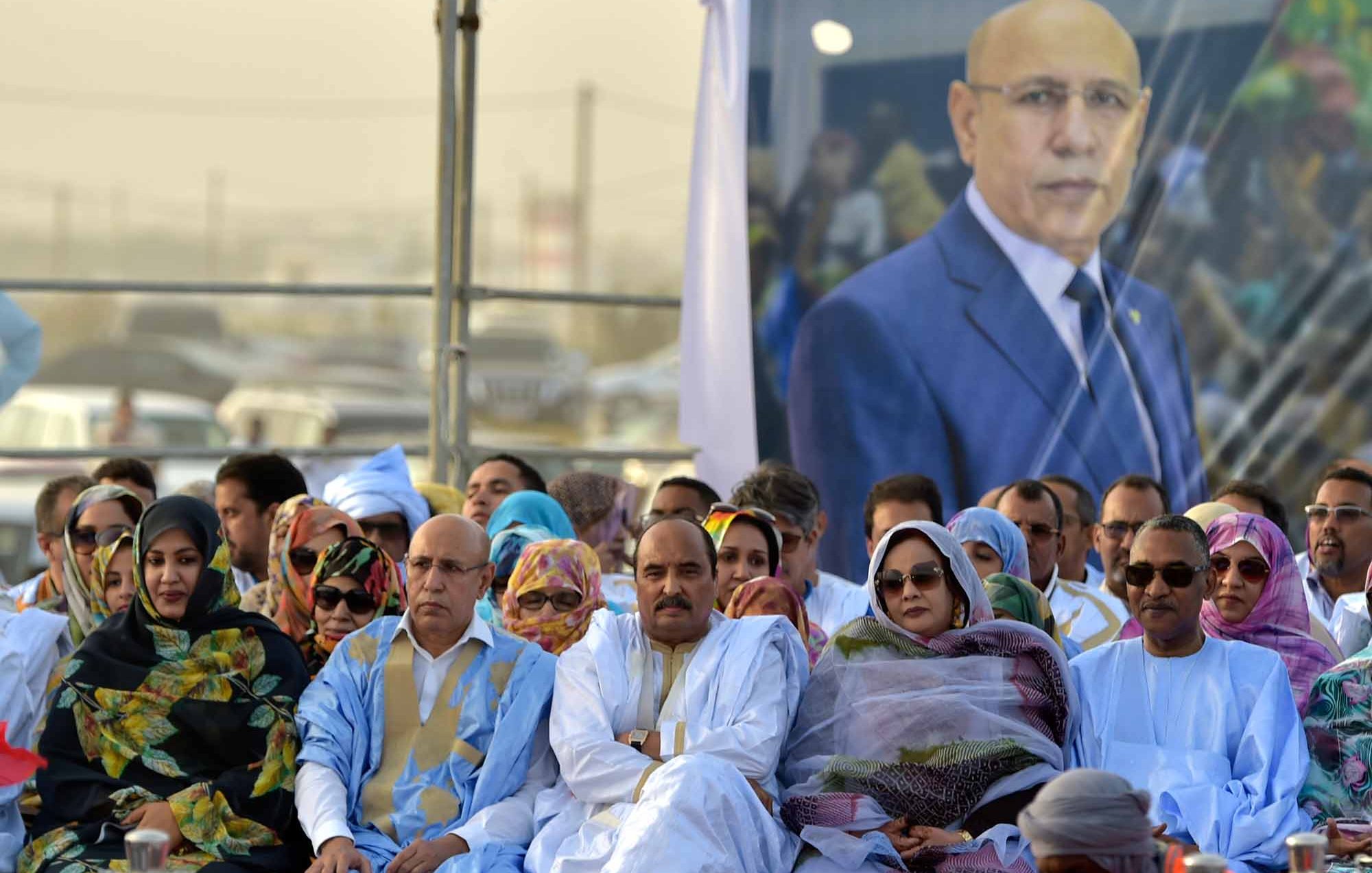 L'image montre un groupe de personnes assises, participant à un événement ou à une cérémonie. Elles portent des vêtements traditionnels, souvent des robes colorées et des turbans. En arrière-plan, il y a un grand portrait d'un homme en costume, probablement une figure politique, qui semble surveiller l'événement. L'ambiance paraît solennelle et impliquée, avec beaucoup d'attention portée au discours ou à la célébration en cours.