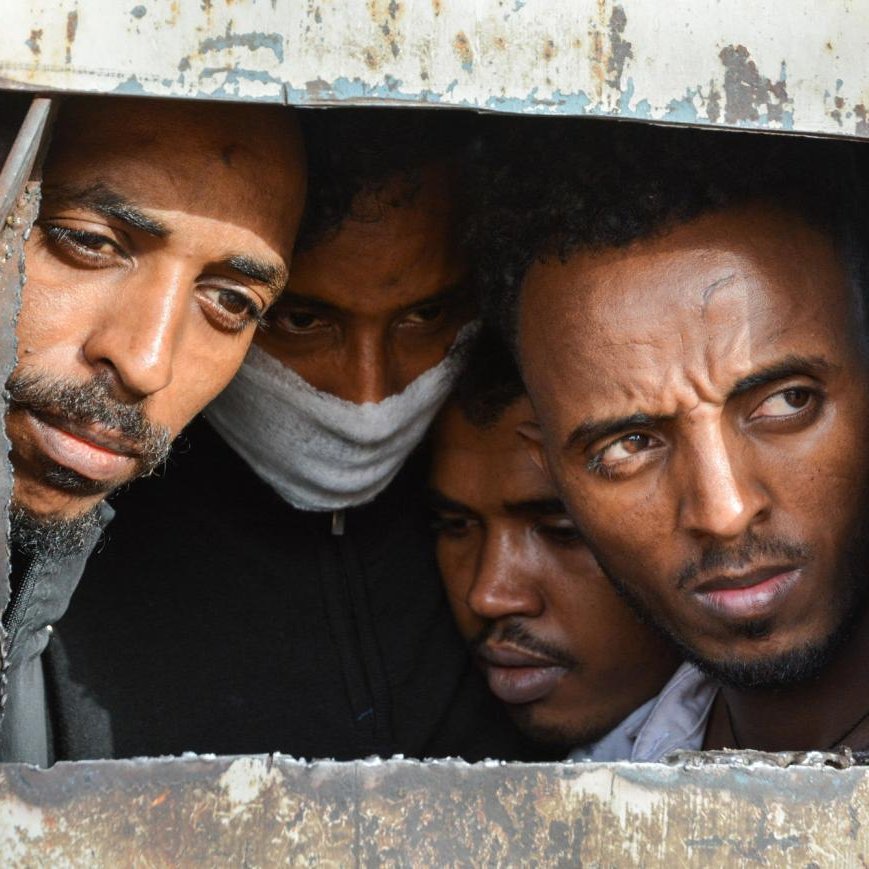 L'image montre plusieurs hommes regardant à travers une petite ouverture, probablement dans un conteneur ou un véhicule. Leur expression semble inquiète ou curieuse, et on peut voir des détails de leur visage, tels que la concentration et une certaine tension. L'arrière-plan est en métal rouillé, ce qui donne une impression de confinement. Les conditions semblent difficiles et l'atmosphère est chargée d'émotion.