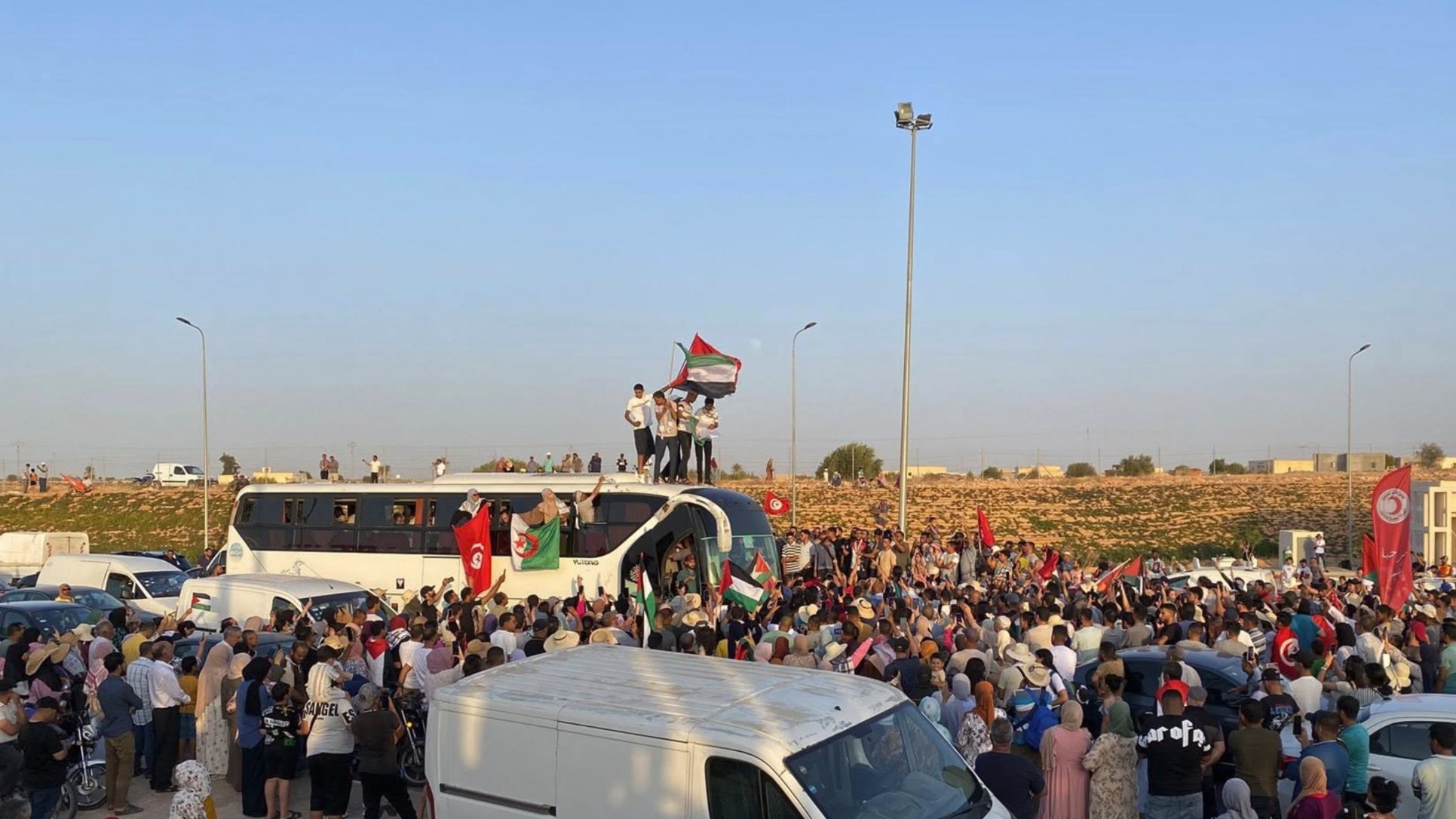 A large crowd gathers, waving flags and celebrating, with vehicles parked around.
