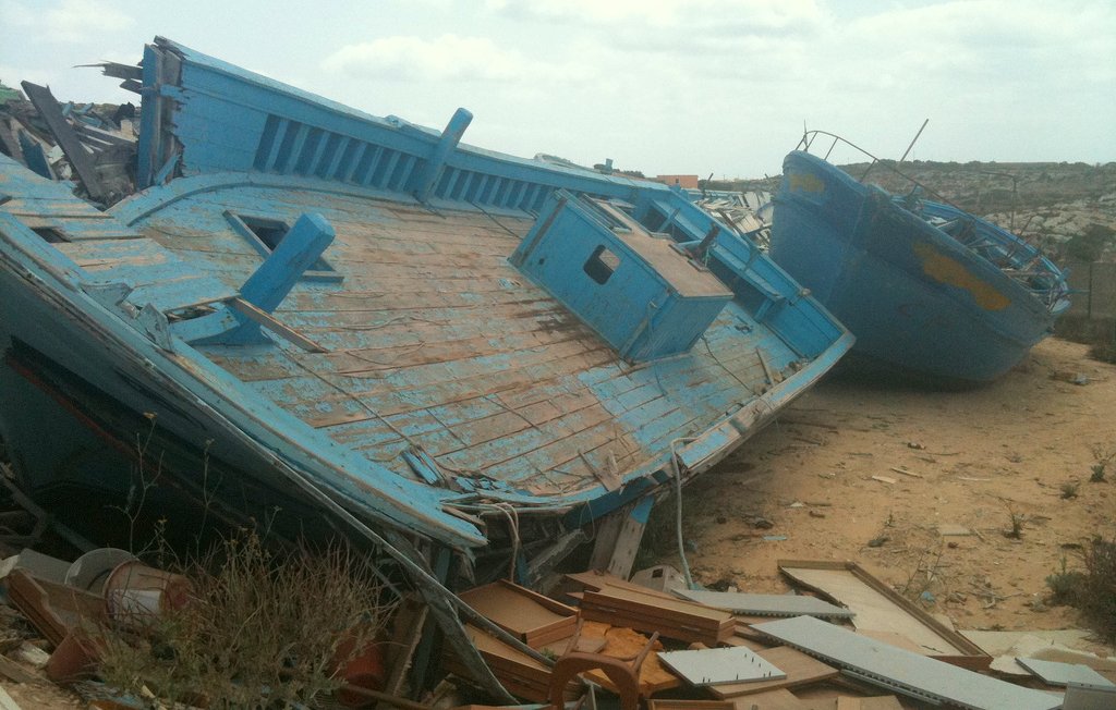 L'image montre des bateaux endommagés, apparemment abandonnés sur une terre aride. Les bateaux, de couleur bleu clair, sont inclinés sur le côté, avec des parties de leur structure exposées. On peut voir des débris et des objets éparpillés autour, ainsi qu'une végétation rudimentaire qui pousse dans le sol. Le ciel est nuageux, créant une atmosphère mélancolique. L'ensemble dégage une impression de désolation et d'abandon.