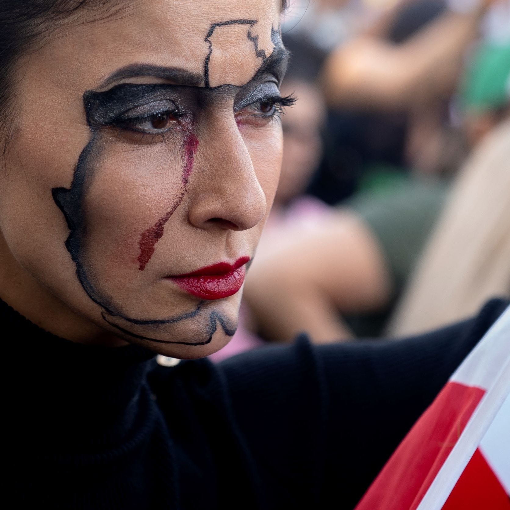 The image features a woman with striking face makeup that outlines the map of a country, highlighting it with dark lines. She has a tear streaking down her cheek, adding an emotional touch to her expression. The woman holds a flag that includes green, white, and red colors, which likely represents her national identity. The setting indicates a public gathering or protest, as there are people in the background, contributing to a vibrant atmosphere.