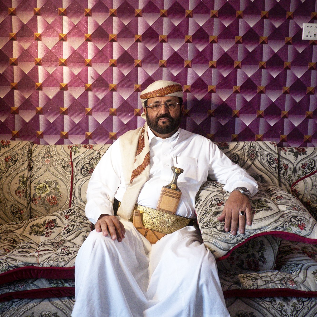 L'image montre un homme vêtu d'un habit traditionnel, assis sur un canapé orné de motifs décoratifs. Il porte une ceinture dans laquelle est insérée une sorte de poignard, typique de certaines cultures arabes. Le fond est constitué d'un mur avec un revêtement texturé en violet, ajoutant une touche colorée à la scène. L'homme a une expression calme et posée, et son environnement reflète une atmosphère accueillante et traditionnelle.