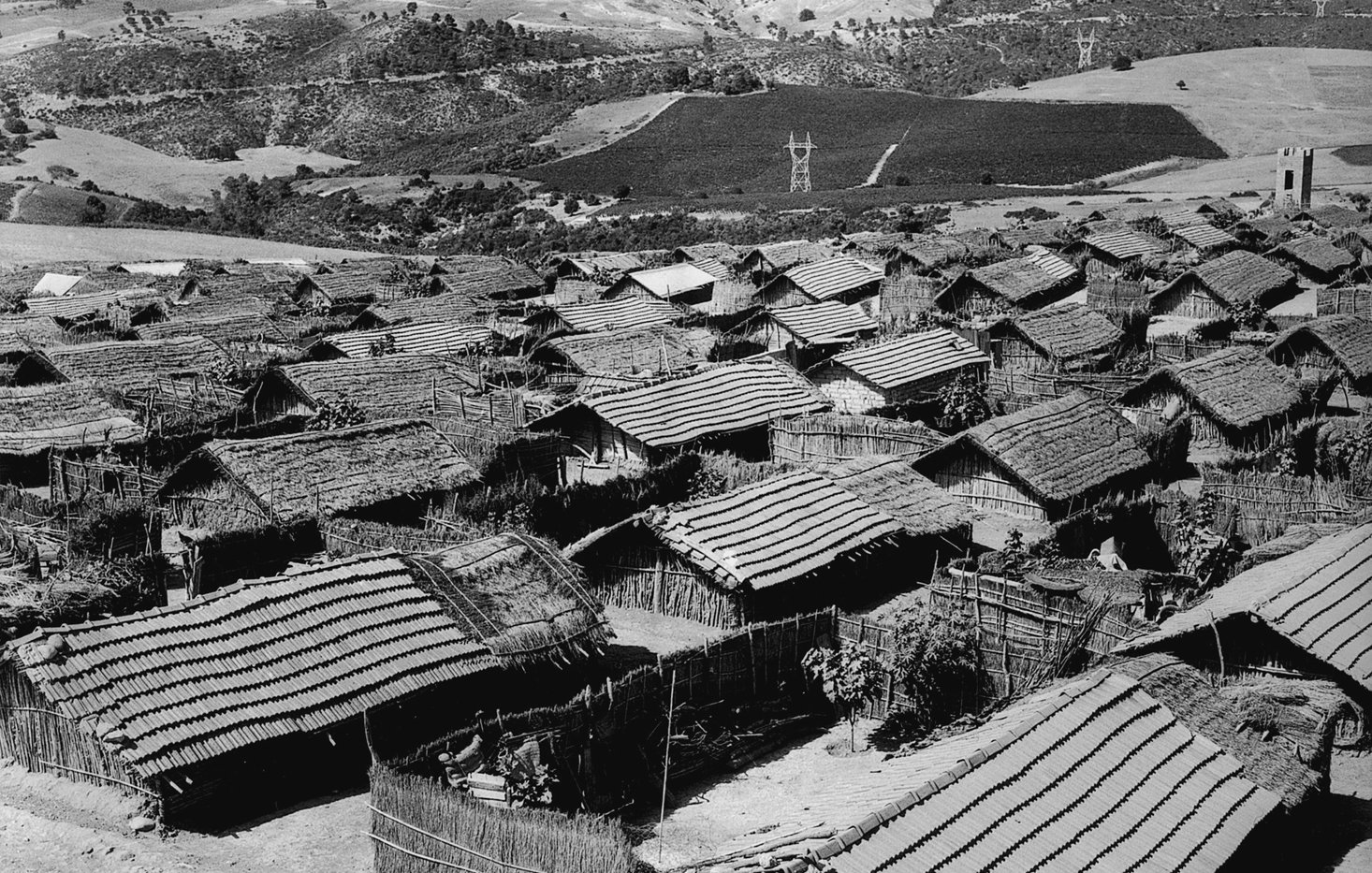 الصورة تُظهر منظرًا عامًّا لقرية صغيرة تتكون من مجموعة من المنازل التقليدية. المنازل تبدو مصنوعة من مواد طبيعية مثل الخشب والقش، مع أسطح مائلة. يتواجد حول القرية تلال خضراء ومناظر طبيعية جميلة، مما يضفي على المشهد طابعًا ريفيًا هادئًا. السماء تبدو صافية، مما يبرز جمال المكان.