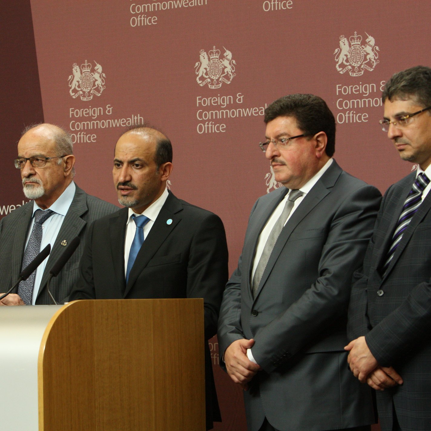 L'image montre un groupe de plusieurs personnes lors d'une conférence de presse. Elles se tiennent devant un podium avec un emblème officiel, et une toile de fond affichant "Foreign & Commonwealth Office". Les individus paraissent sérieux et concentrés, probablement en train de faire une déclaration ou de répondre à des questions. Les personnes sont habillées de manière formelle, illustrant l'importance de l'événement.