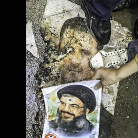 L'image montre un sol recouvert de débris, avec un visage décoloré visible sur le sol. Une personne porte une chaussure qui marche sur une partie de l'image, tandis qu'une autre main tient un poster montrant un homme en souriant, probablement une figure publique. Le tout évoque un contexte de manifestation ou de désapprobation.