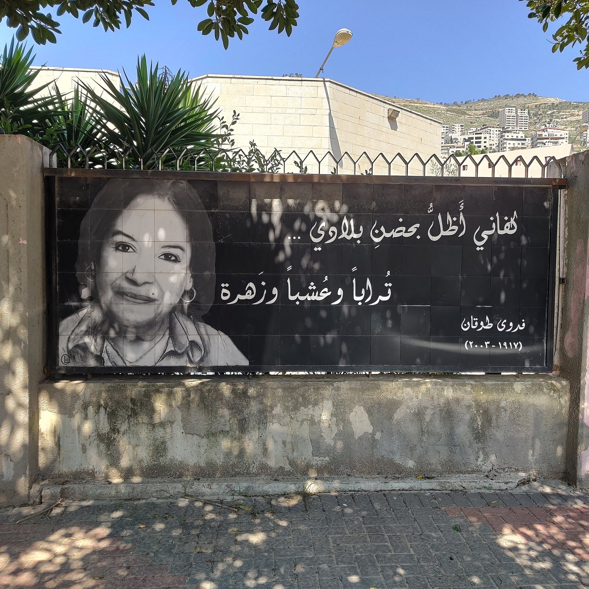 L'image montre une grande affiche accrochée à une clôture. Elle présente un portrait en noir et blanc d'une femme souriante. À côté de son visage, on trouve une citation en arabe, ainsi qu'un nom et une date en bas. L'arrière-plan montre des bâtiments et des végétaux, indiquant que l'affiche est placée dans un environnement urbain. L'ensemble véhicule une ambiance d'hommage ou de souvenir.