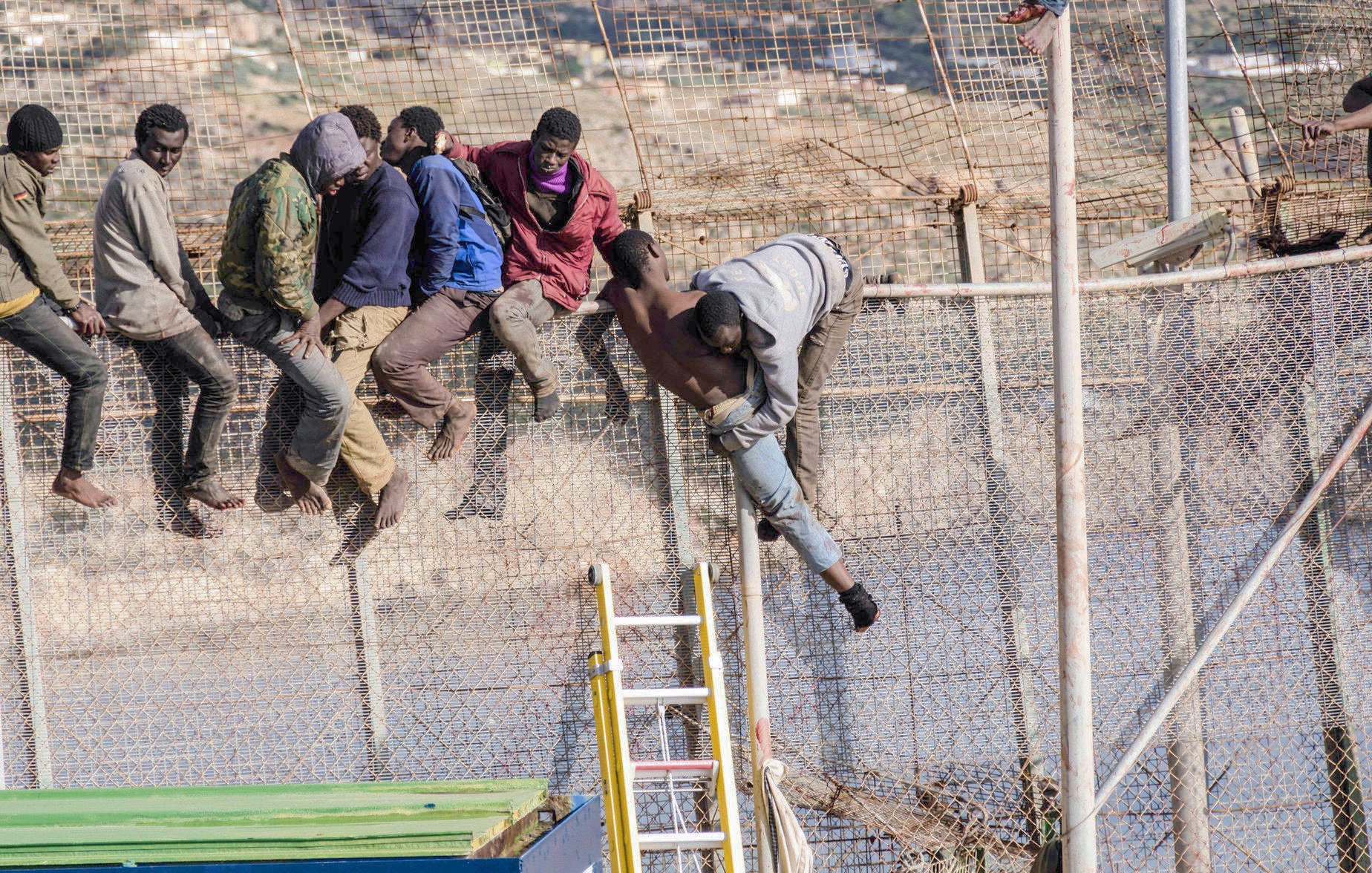 L'image représente un groupe de personnes tentant de franchir une clôture métallique. On peut voir plusieurs individus en train de grimper et de passer par-dessus la barrière, certains sont déjà en train de descendre de l'autre côté. L'environnement semble être une zone frontalière, probablement dans un contexte de migration. Les personnes semblent déterminées, et l'image transmet une forte impression d'urgence et de mouvement.