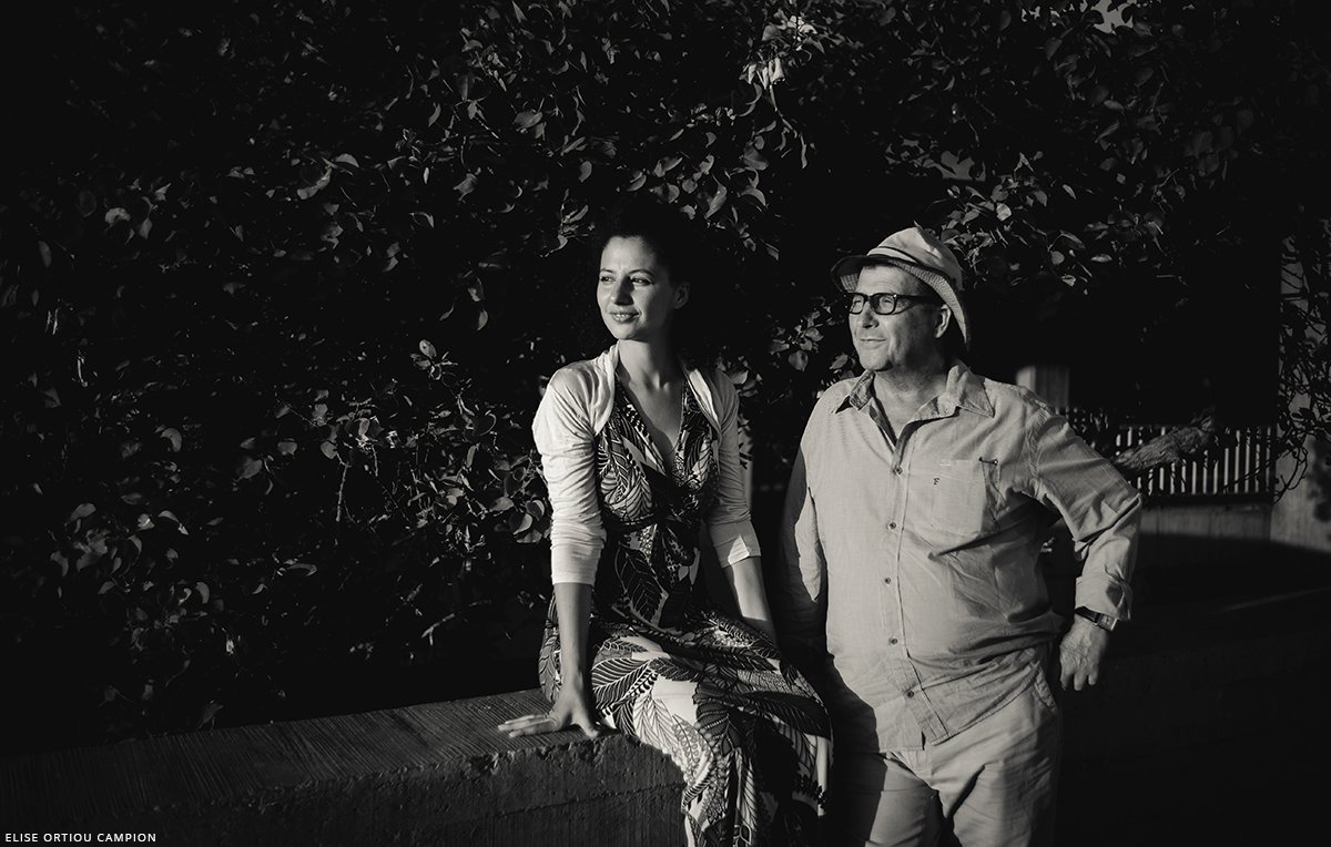 L'image montre une femme et un homme, tous deux dans un cadre extérieur, probablement près d'une plante ou d'un mur recouvert de verdure. La femme, habillée d'une robe imprimée, se tient assise sur un muret avec une posture détendue et un regard pensif. L'homme, vêtu d'un chemisier léger et d'un chapeau, se tient à côté d'elle avec un sourire, ajoutant à l'atmosphère conviviale de la scène. Le contraste entre les ombres et la lumière crée une ambiance artistique et intemporelle, en noir et blanc.