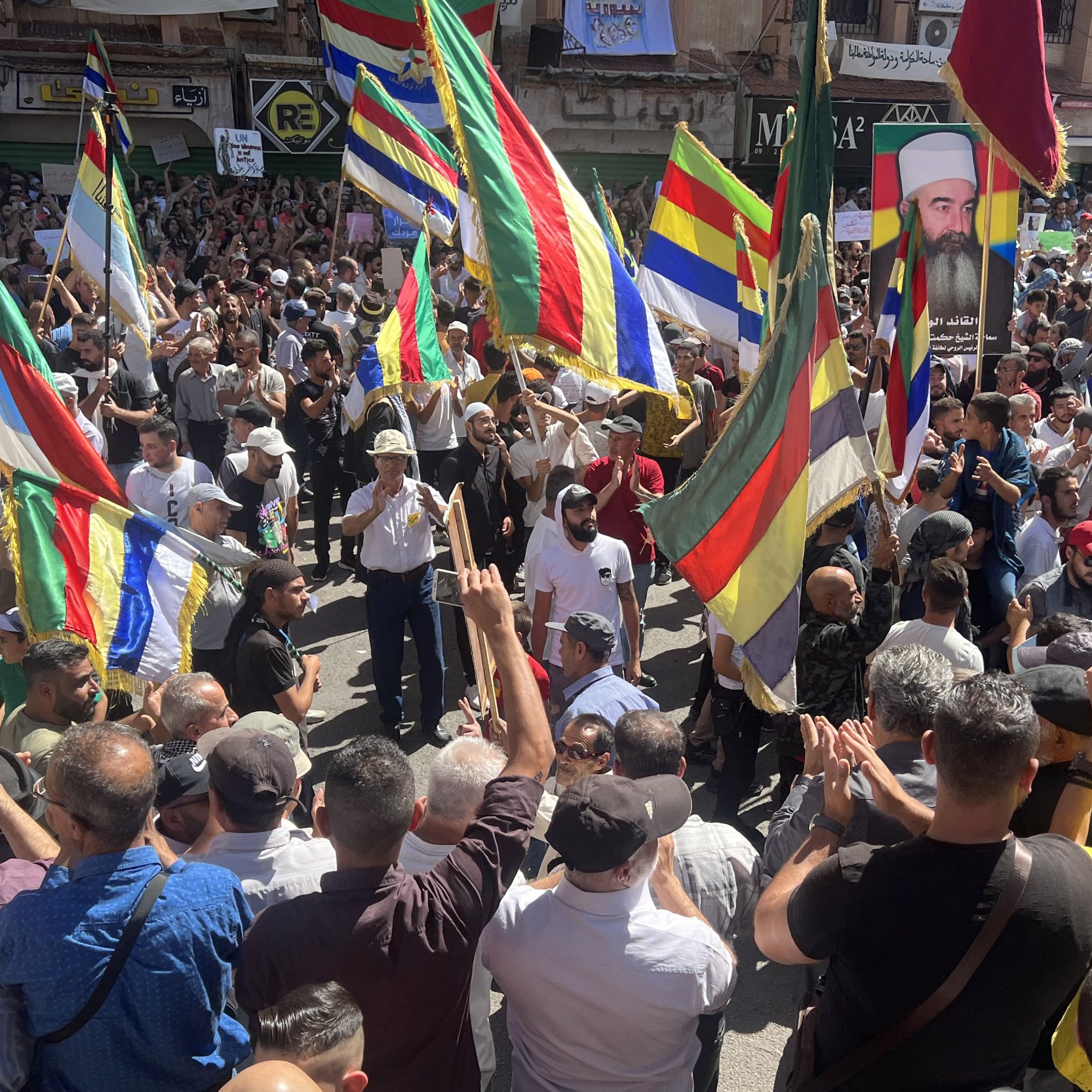 The image depicts a large crowd gathered in a public space, participating in a demonstration or protest. Many people are holding brightly colored flags that feature a mix of horizontal and vertical stripes. The atmosphere appears energetic and spirited, with participants actively engaging with one another. Some individuals are raising their hands and clapping, suggesting a celebratory or passionate mood. In the background, various signs and posters can be seen, indicating that the event is likely focused on a specific cause or issue.