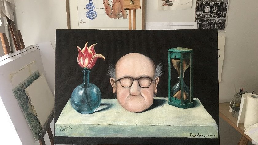 L'image montre une toile peinte, représentant un homme avec une tête large, des lunettes et des cheveux ébouriffés. Il est placé devant une table sur laquelle se trouvent un vase avec une fleur rouge, une sablier et un autre objet qui semble être une bouteille. En arrière-plan, on aperçoit des œuvres d'art accrochées au mur, ainsi qu'un chevalet. L'ensemble a une atmosphère artistique et quelque peu surréaliste.