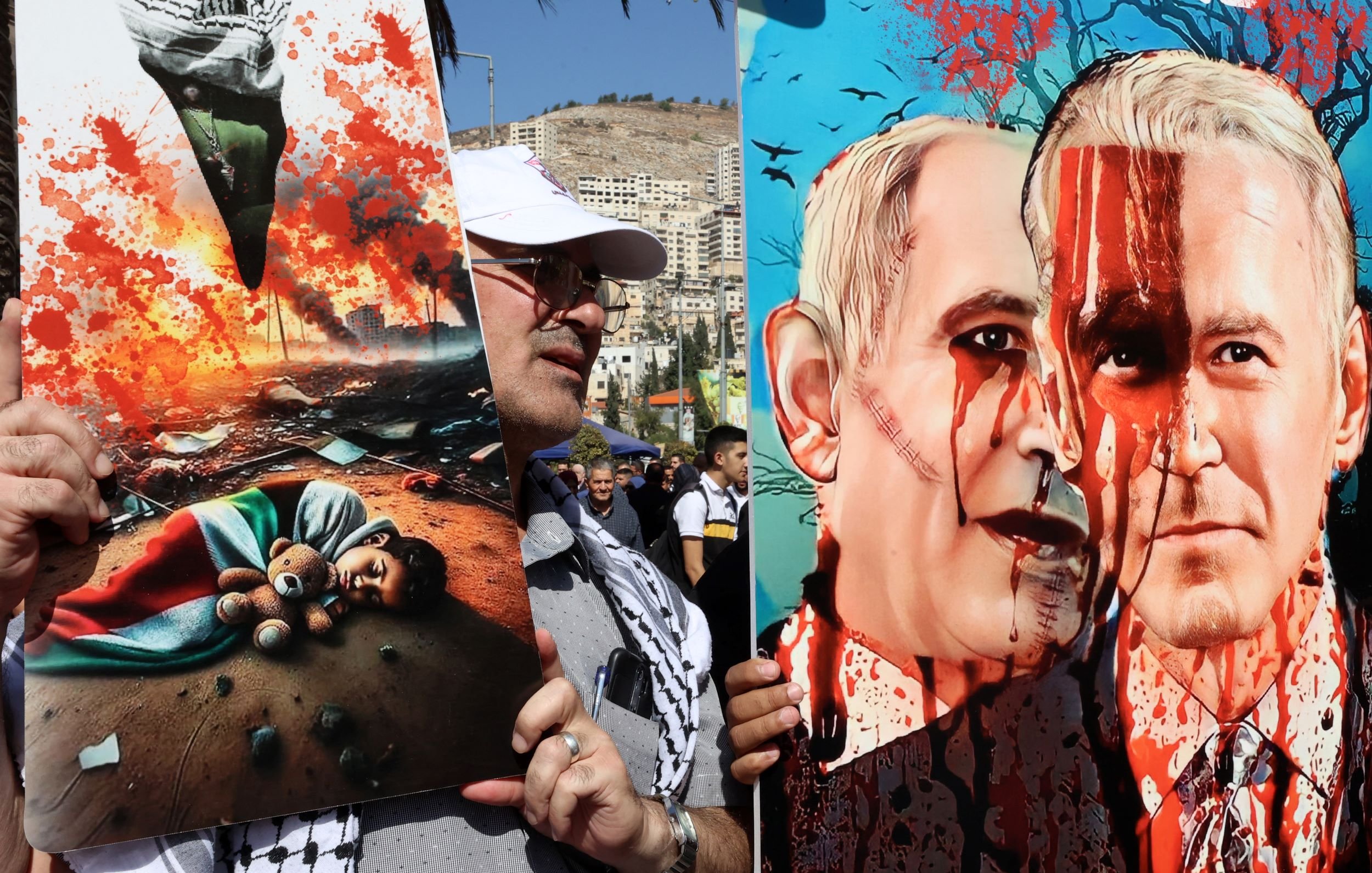 The image features two striking pieces of protest art held by a person. On the left, there is a depiction of a devastated scene showing a child's lifeless body and a teddy bear amidst destruction, conveying a powerful message about the impact of conflict on innocent lives. On the right, another artwork portrays two figures resembling political leaders, with exaggerated features and blood splatters, suggesting themes of violence and political aggression. The overall mood of the image is one of protest and strong emotional expression regarding war and its consequences.