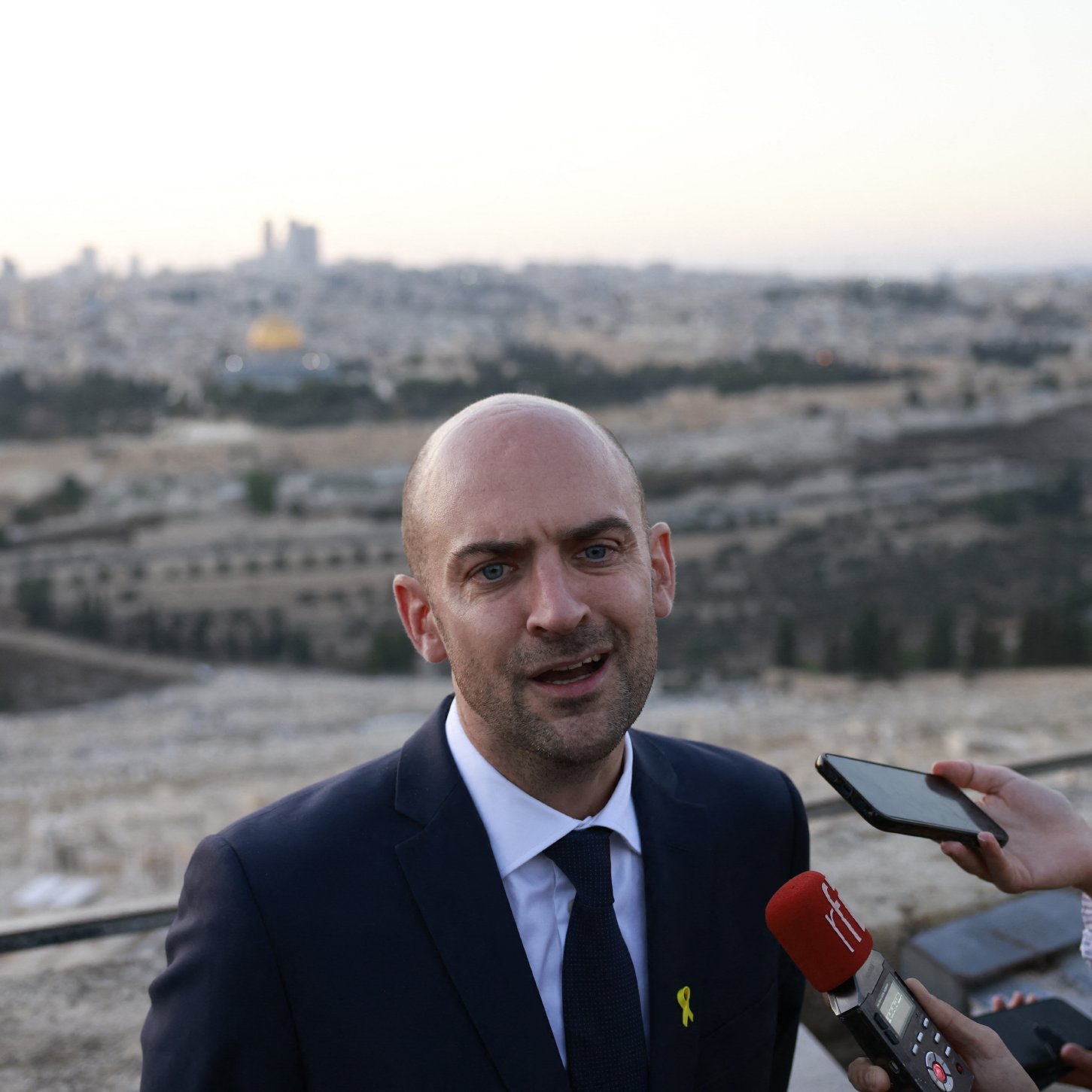 در این تصویر، یک مرد با سر طاس و کت و شلوار تیره در حال صحبت به دوربین و خبرنگاران است. او در یک مکان آزاد ایستاده و پشتش چشم‌انداز شهر قابل مشاهده است. درختان و ساختمان‌ها در دوردست دیده می‌شوند، و به نظر می‌رسد که این تصویر در یک زمان غروب یا صبح زود گرفته شده است. خبرنگاری نیز در سمت راست او قرار دارد و در حال ثبت‌کردن یا پرسش کردن از اوست.