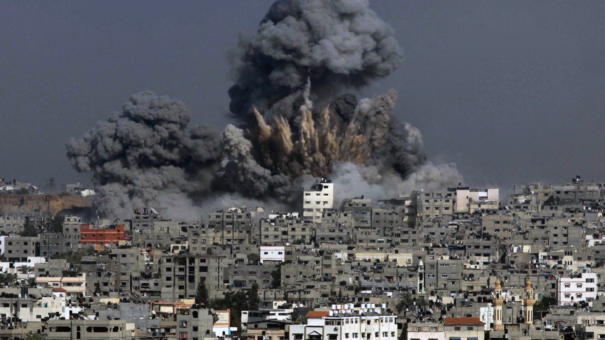 The image depicts a scene of destruction with a large explosion occurring in an urban area. Thick, dark gray smoke billows into the sky, rising above the buildings in the foreground. The skyline is dominated by closely packed structures, showing signs of damage. The overall atmosphere conveys a sense of chaos and devastation, characteristic of an area affected by conflict or military action.