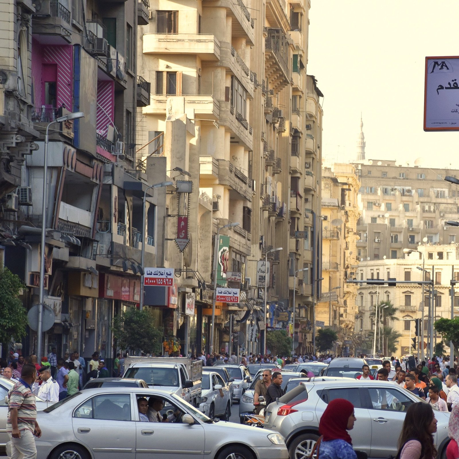 L'image montre une rue animée remplie de voitures et de passants. On peut voir des bâtiments de plusieurs étages de part et d'autre de la rue, avec des balcons et des façades variées. La scène semble se dérouler pendant la journée, la lumière du soleil illuminant l'environnement urbain. Des panneaux de signalisation et des enseignes en arabe sont visibles, ajoutant à l'atmosphère locale de cette ville. Les gens semblent se déplacer rapidement, créant une ambiance dynamique et vivante.