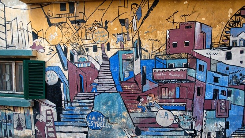 L'image montre une murale colorée peinte sur un mur, représentant une scène de la vie urbaine. On y voit des escaliers, des maisons aux façades vives, et des personnages en train de vaquer à diverses activités. La murale semble évoquer la culture et l'esprit d'une communauté, avec des mots tels que "LA VIE" et "ESBELLE" intégrés dans le dessin. Les couleurs utilisées sont vives et variées, ajoutant une ambiance dynamique et joyeuse à la scène.