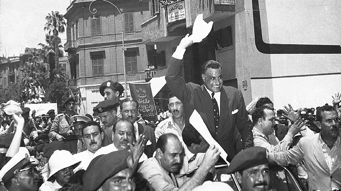 L'image montre une scène de foule animée, probablement lors d'un événement historique. De nombreuses personnes sont rassemblées autour d'une voiture, où un homme, visiblement en position d'autorité, lève les bras en signe de salutation ou d'approbation. Les spectateurs semblent enthousiastes, agitant des banderoles ou des papiers. On peut voir des policiers et des militaires en uniforme, ajoutant à l'ambiance de la scène. L'architecture des bâtiments en arrière-plan suggère une ville dynamique, probablement dans un contexte politique ou social significatif.