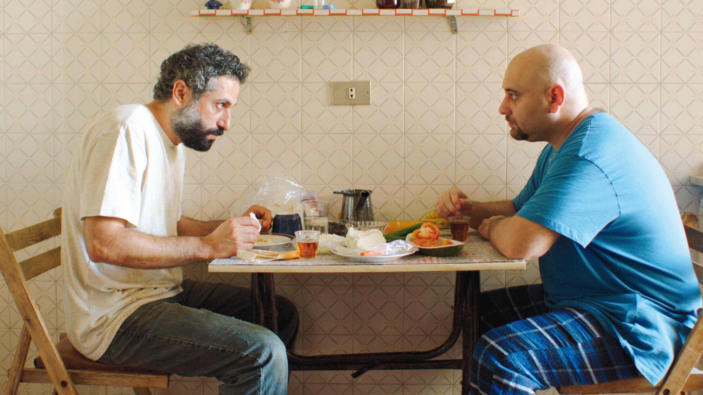 L'image montre deux hommes assis à une table dans une pièce au décor simple. Ils semblent en pleine conversation, chacun portant une expression sérieuse. Sur la table, il y a des assiettes, des verres et des aliments. L'un des hommes porte un t-shirt gris, tandis que l'autre est vêtu d'un t-shirt bleu. La pièce est éclairée de façon naturelle, et le mur est décoré d'un motif en damier. L'ambiance semble à la fois intime et chargée.