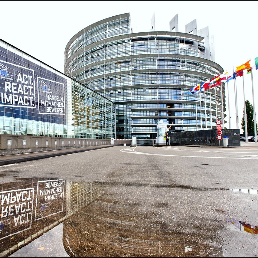 L'image représente un bâtiment moderne avec une architecture distinctive, probablement un siège d'institution européenne. On peut voir des fenêtres en verre réfléchissant des phrases comme "AGIR, RÉAGIR, ACCOMPLIR" en plusieurs langues. À droite, il y a une série de drapeaux représentant différents pays, tandis qu'une flaque d'eau au premier plan reflète l'édifice et les messages affichés sur la façade. L'ensemble donne une impression de dynamisme et de modernité.
