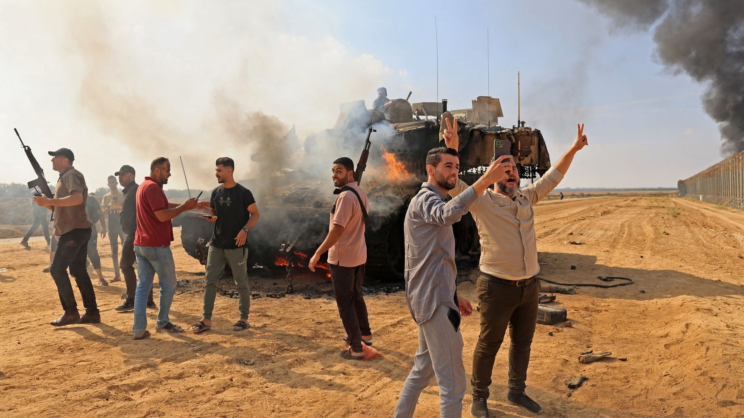 La imagen muestra un grupo de hombres en un entorno de conflicto. Están en un terreno árido y polvoriento, con humo negro elevándose en el fondo, lo que sugiere actividad bélica. Algunos de los hombres están armados y parecen expresar una mezcla de emociones, como alegría o desafío, mientras hacen gestos con las manos. También hay un vehículo militar dañado en el centro de la escena, lo que indica que se ha producido un enfrentamiento.