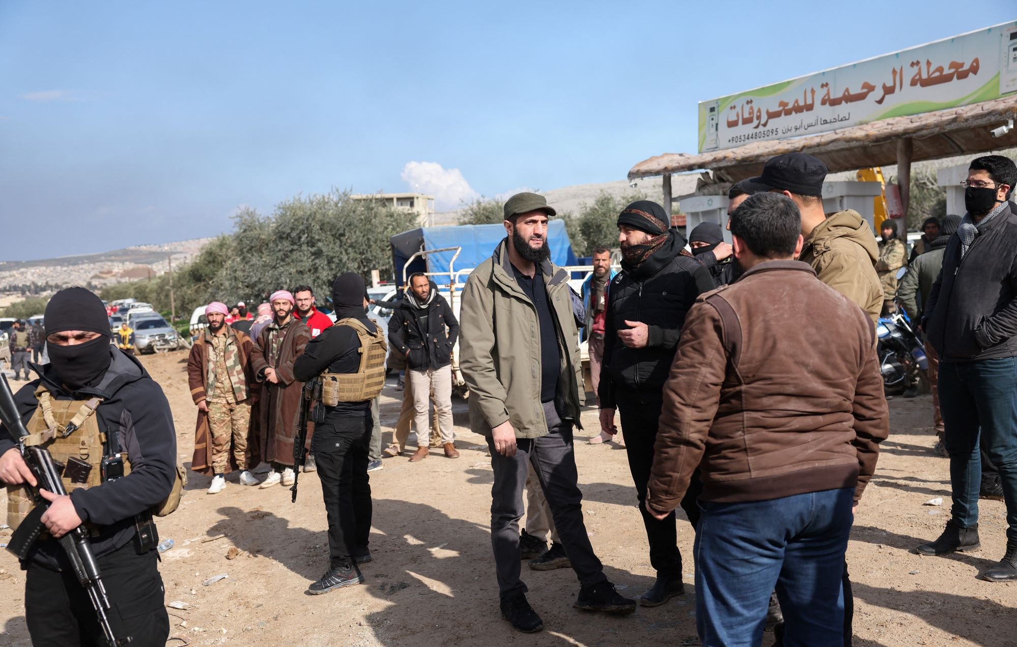 تظهر الصورة مجموعة من الأشخاص في منطقة مفتوحة تحت سماء زرقاء مع بعض الغيوم. يبدو أن هناك عددًا من الرجال يرتدون ملابس تقليدية وبعضهم يرتدي ملابس عسكرية أو شبه عسكرية. في الخلفية، يوجد لافتة تشير إلى "محطة الرحمة للمشاريع". يُلاحظ أن بعض الأشخاص يتحدثون مع بعضهم البعض، وهناك أيضًا مجموعة من الأشخاص يحملون أسلحة. الأجواء تبدو نشطة وتعبر عن تفاعل اجتماعي بين الحضور.