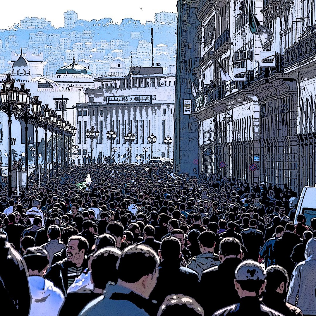 L'image montre une foule dense de personnes marchant dans une rue, probablement dans une ville animée. Les bâtiments sur les côtés semblent historiques, avec une architecture riche. On peut voir des lampadaires le long de la promenade, et au loin, on aperçoit des collines ou des montagnes qui ajoutent à la profondeur de la scène. L'atmosphère semble vibrante et dynamique, reflétant probablement une manifestation ou un événement public.