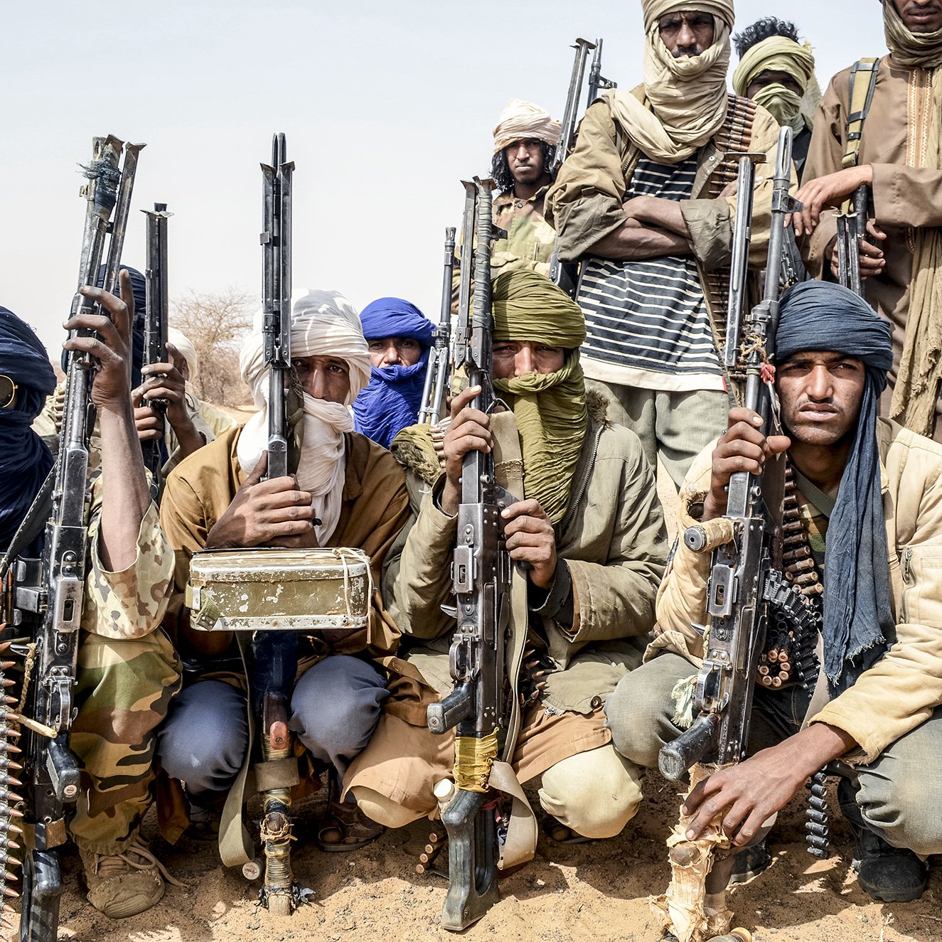 L'image montre un groupe d'hommes armés, probablement dans un environnement désertique. Ils portent des vêtements typiques de régions arides, comme des turbans et des foulards qui cachent partiellement leur visage. Chaque homme tient une arme à feu, et l'ambiance générale semble sérieuse et déterminée. Le groupe est organisé et se présente en ligne, ce qui suggère une certaine discipline. Le fond désertique renforce la dureté du cadre dans lequel ils se trouvent.