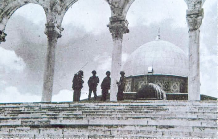L'image montre une scène en noir et blanc, probablement historique, avec un groupe de soldats se tenant sur des marches devant un édifice. On peut apercevoir un dôme caractéristique au fond, ainsi que des arches en pierre en avant-plan. L'atmosphère est solennelle et le lieu semble chargé d'histoire. Les soldats sont habillés de manière militaire et semblent en position de garde.