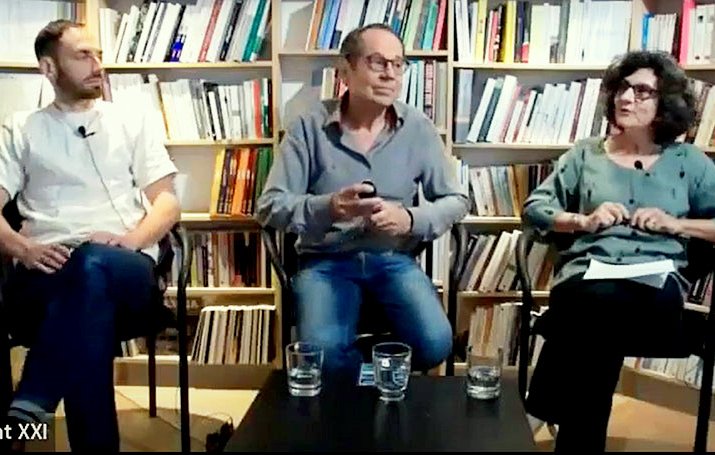 Cette image montre trois personnes assises sur des chaises dans un environnement intérieur, probablement une librairie ou un espace de discussion. À l'arrière-plan, il y a des étagères remplies de livres. La personne à gauche porte une chemise blanche, celle du centre est en chemise grise et celle à droite porte un haut vert. Ils semblent être en train de discuter ou de débattre, avec des verres d'eau devant eux. L'ambiance semble détendue et propice à l'échange d'idées.