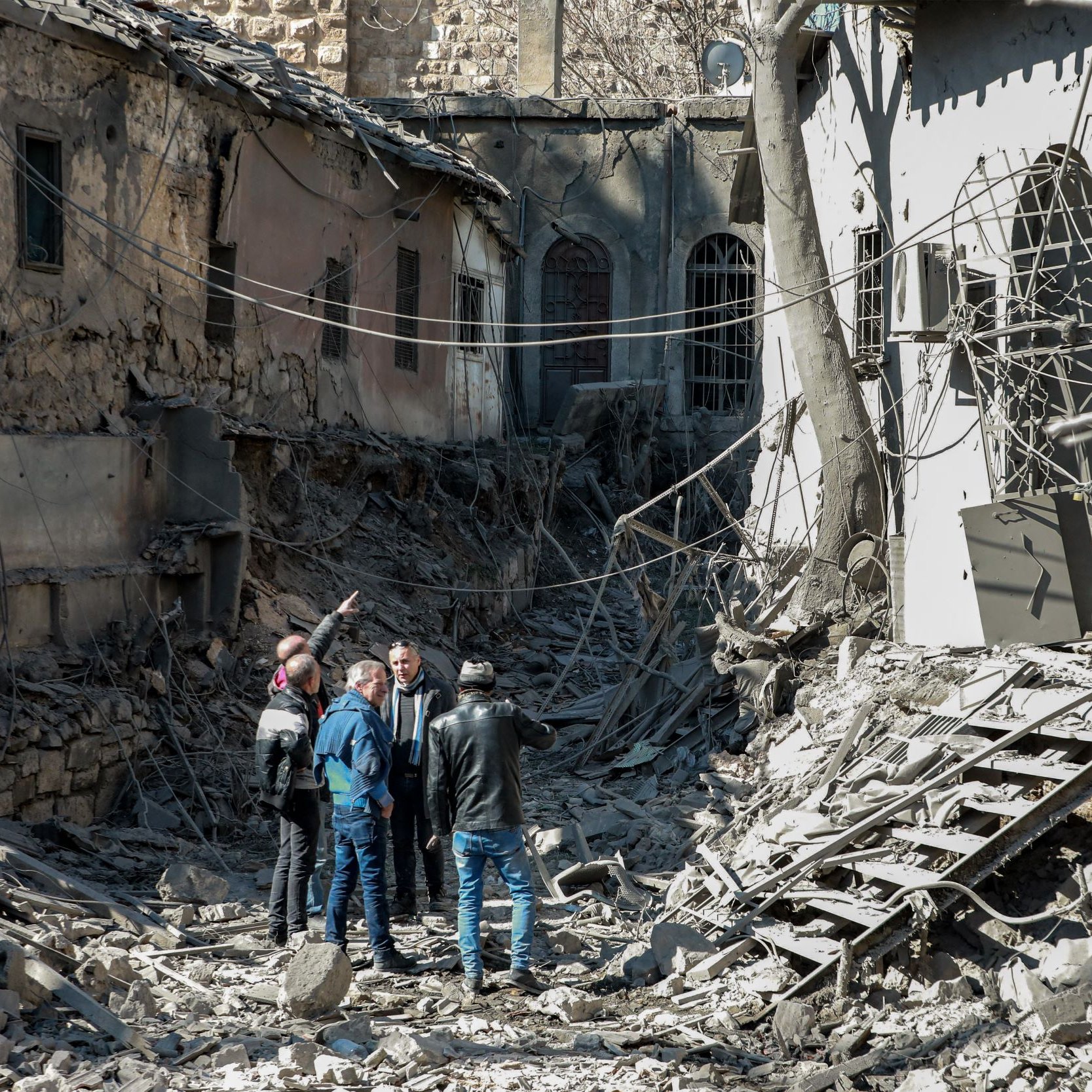 L'image montre un groupe de personnes se tenant au milieu d'un paysage de destruction. Les bâtiments autour d'eux sont en ruines, avec des murs effondrés et des débris éparpillés sur le sol. Les personnes semblent discuter ou examiner les lieux, tandis que des fils électriques sont visibles dans l'environnement. L'atmosphère est marquée par un sentiment de désolation et de perte.