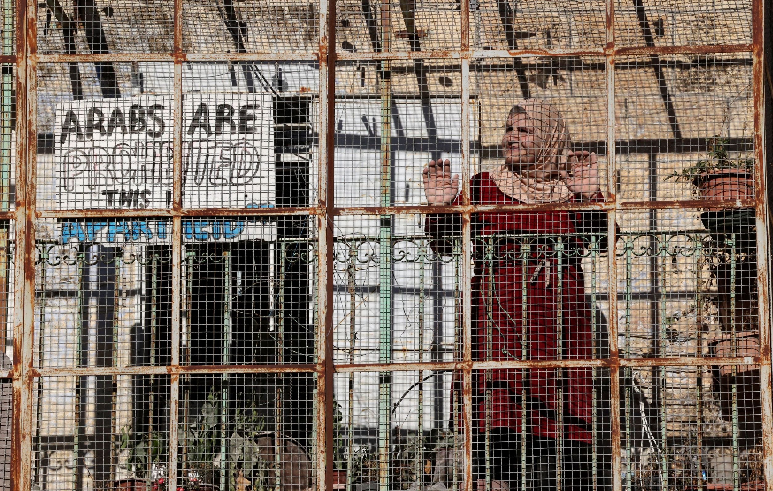 L'image montre une femme se tenant derrière une structure en métal grillagée. Elle porte un habit traditionnel et semble regarder à travers les barreaux. En arrière-plan, on aperçoit un panneau avec des mots en anglais qui semblent exprimer un message, mais son contenu exact reste obscurci par la grille. La scène évoque des thèmes de confinement et de séparation, avec une atmosphère de tension ou de défi face à une réalité difficile. Des plantes sont visibles dans l'environnement.