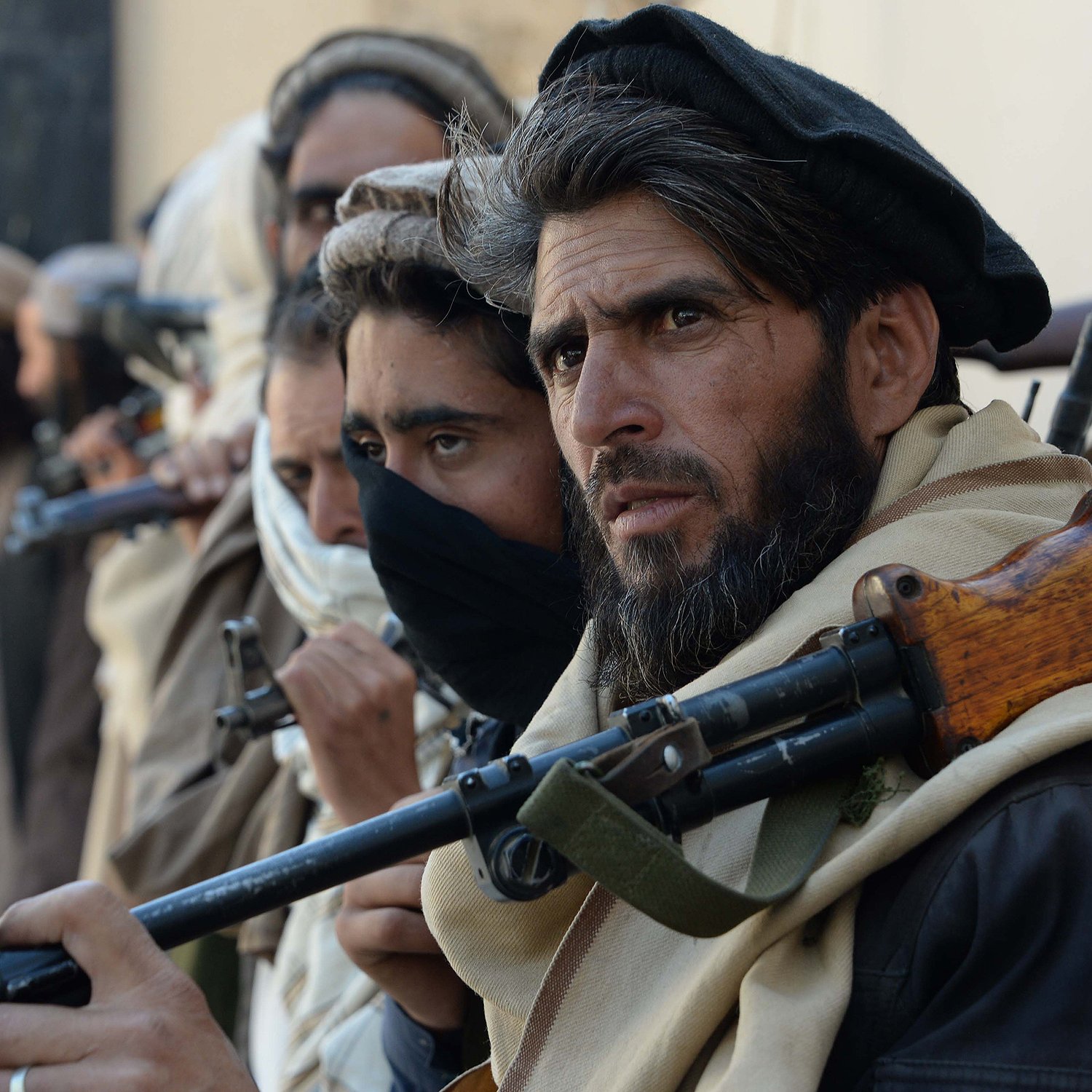 L'image montre un groupe de personnes, apparemment des combattants, qui se tiennent alignés. Ils portent des vêtements traditionnels, certains avec des écharpes ou des masques couvrant leur visage. Chaque individu semble tenir une arme, et leurs expressions sont sérieuses et concentrées. L'environnement semble être urbain ou désertique, avec un fond flou qui suggère une atmosphère de tension.