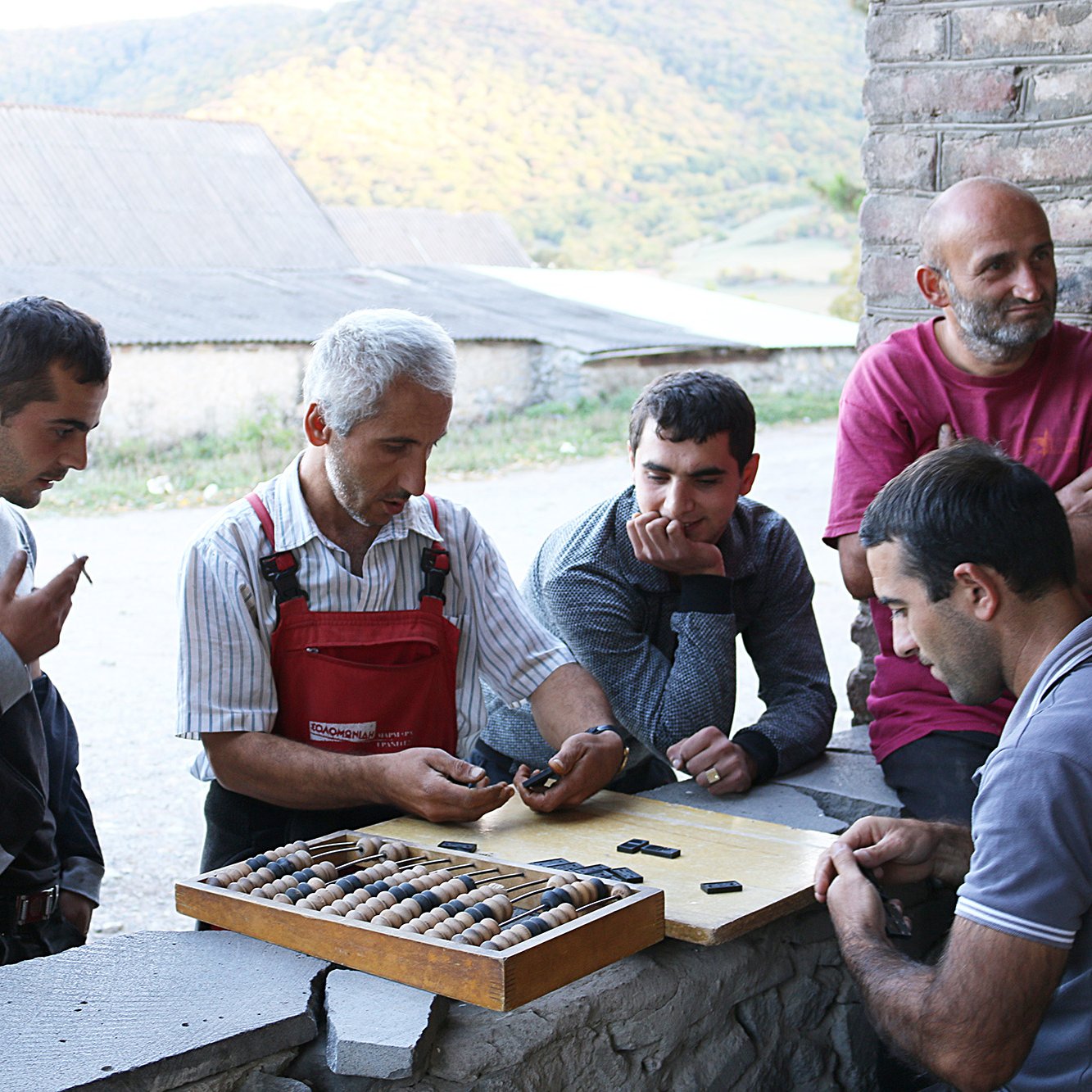 تظهر في الصورة مجموعة من الرجال يجتمعون حول طاولة في مكان يبدو ريفيًا. الرجال الأربعة يبدون مهتمين بشيء ما على الطاولة. أحدهم يحمل عدة أدوات صغيرة، بينما ينظر الآخرون بانتباه. يحيط بهم طبيعة خضراء جميلة، مع تلال في الخلفية. الجو يبدو غير رسمي ومريح، مما يدل على اجتماع اجتماعي أو نشاط جماعي.