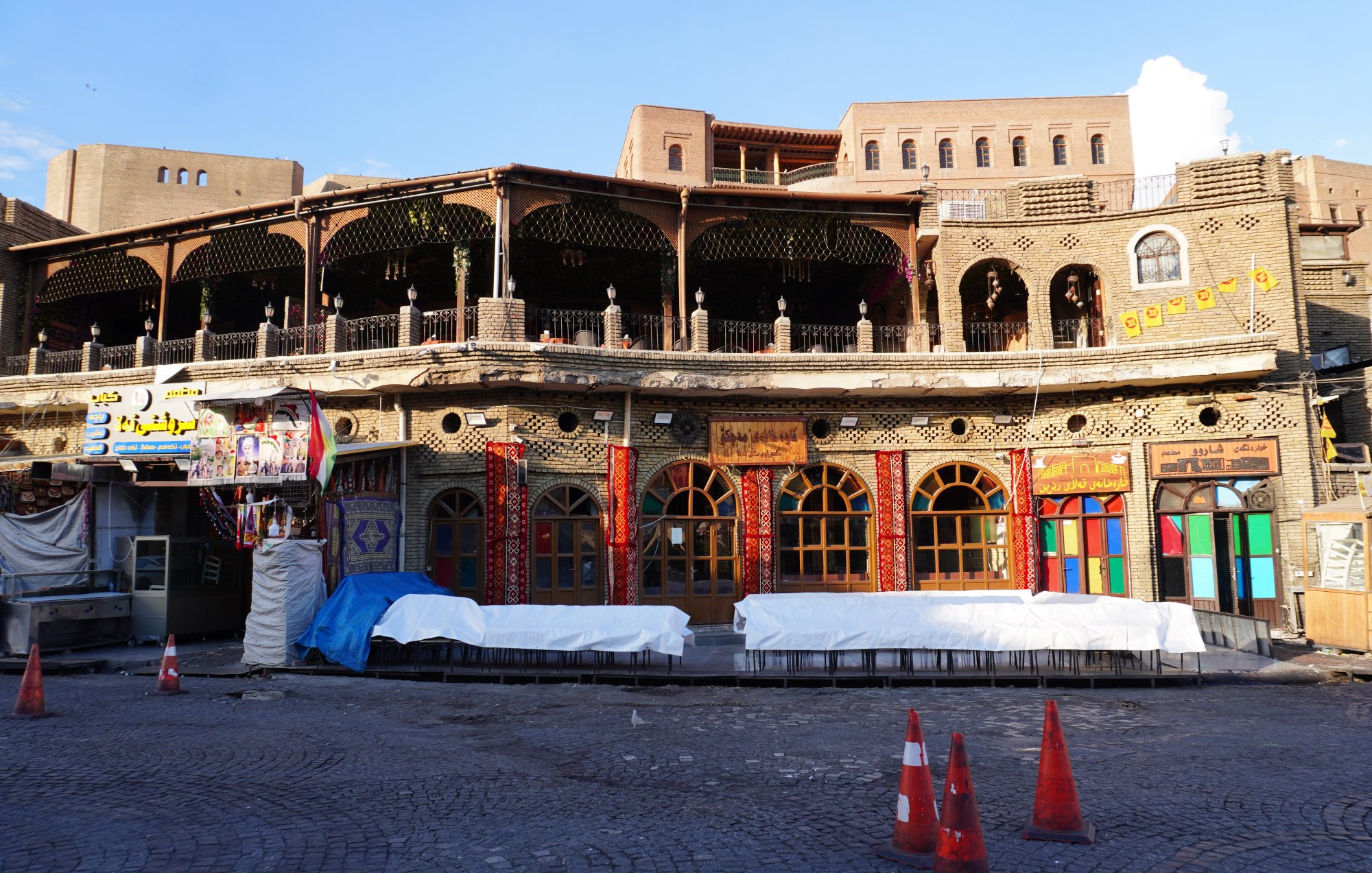 The image depicts a beautifully constructed building featuring intricate architectural details, likely in a historic or culturally significant area. The facade showcases ornate designs, colorful window frames, and a balcony adorned with wooden railings. The ground level appears to have some protective coverings, possibly due to ongoing renovations or maintenance. Surrounding the building, there are orange traffic cones indicating restricted access in that area. The overall ambiance suggests a blend of tradition and vibrant community life.