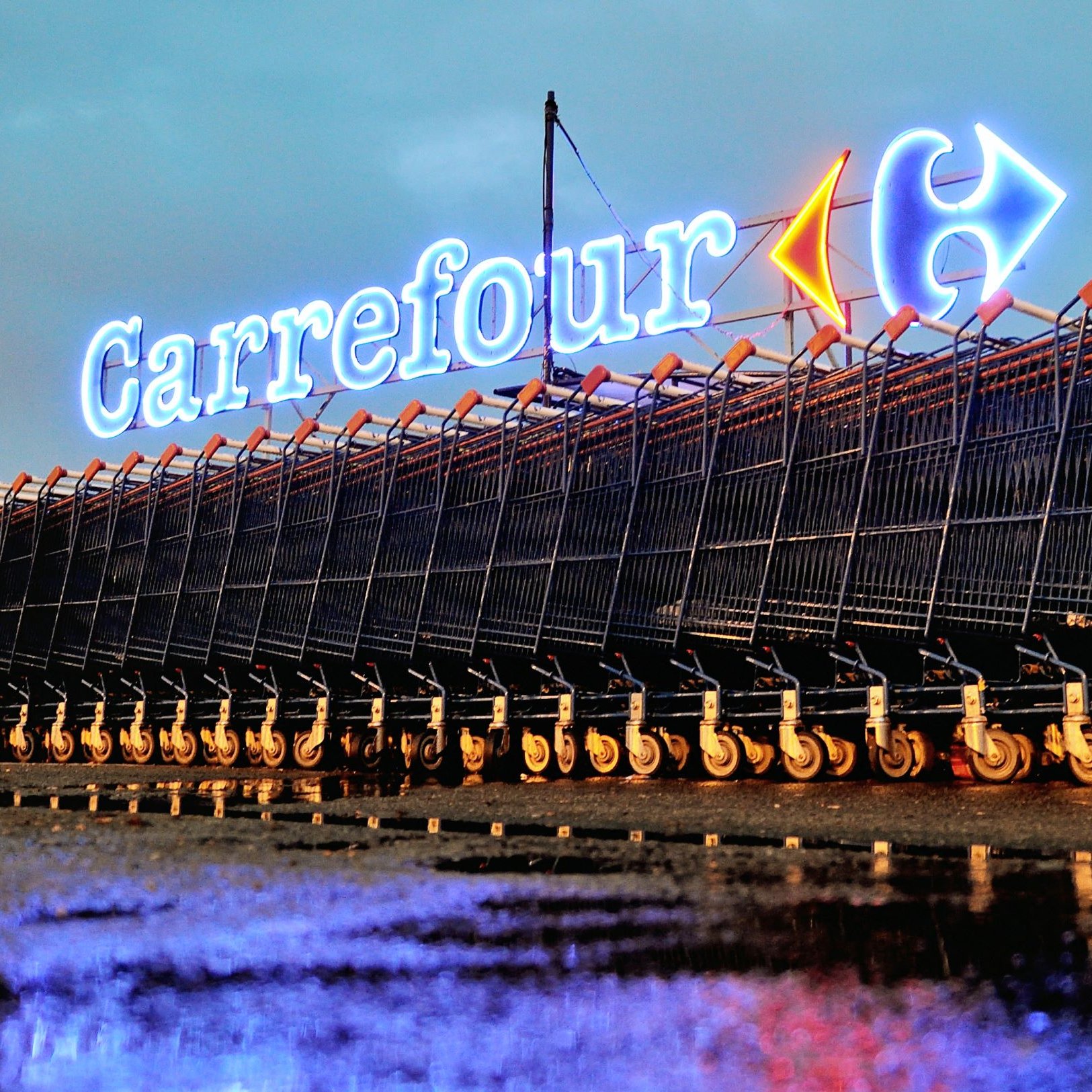 L'image montre un rayonnement lumineux du logo de Carrefour, un hypermarché bien connu, en arrière-plan. Devant le logo, il y a une série de caddies alignés sur le sol, probablement après la pluie, ce qui crée des reflets sur la surface. L'atmosphère est calme et nocturne, accentuée par l'éclairage des enseignes. Les caddies sont organisés de manière ordonnée, offrant une vue intéressante sur l'environnement d'un magasin de grande distribution.