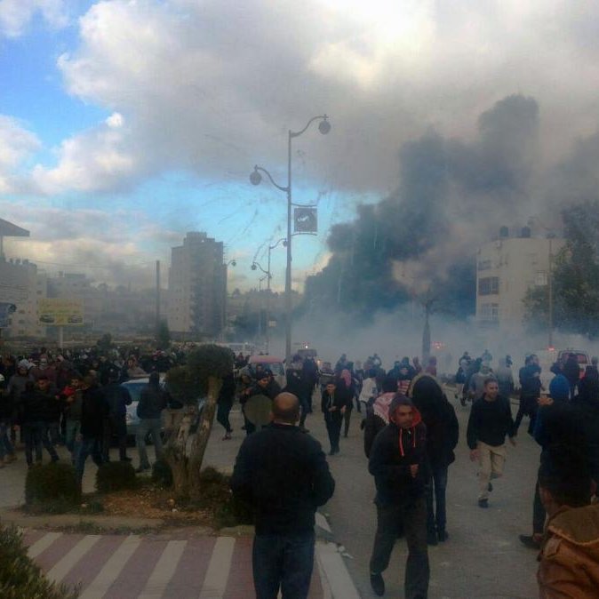 L'image montre une scène de manifestation dans une ville, avec une foule rassemblée dans une rue. Il y a des nuages de fumée et des signes de tension, suggérant des troubles ou des affrontements. Les gens marchent, certains semblent fuir la fumée, tandis que des bâtiments et des arbres sont visibles en arrière-plan. Le ciel est partiellement nuageux, ajoutant à l'atmosphère dramatique de la scène.