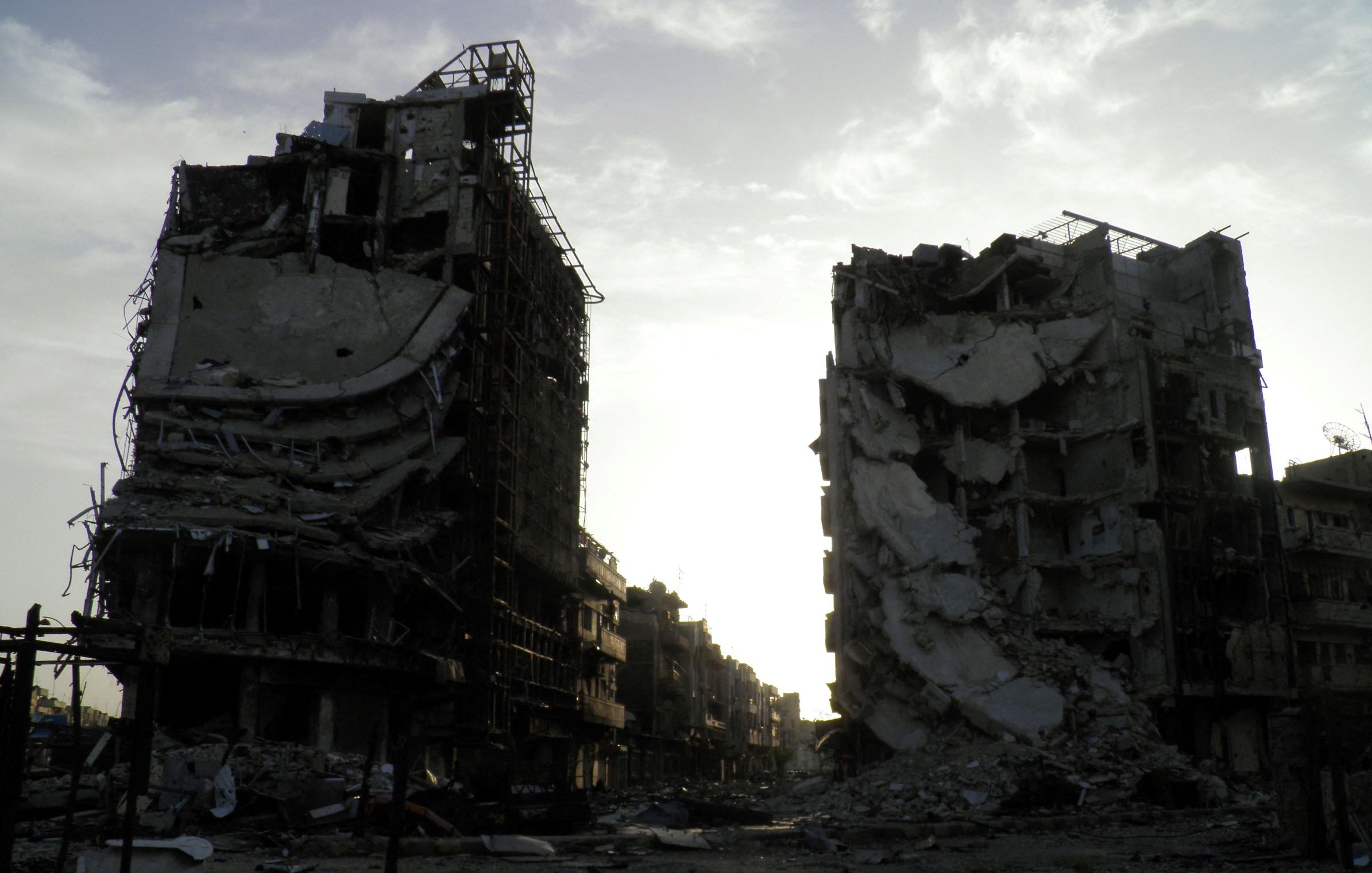 L'image montre deux bâtiments en ruines, témoignant des conséquences d'un conflit ou d'une catastrophe. Les structures sont partiellement effondrées, avec des débris dispersés autour. L'éclairage est sombre, suggérant un crépuscule ou une journée nuageuse, ce qui ajoute une atmosphère de désolation à la scène. Le paysage environnant semble aussi dévasté, accentuant la gravité de la situation.