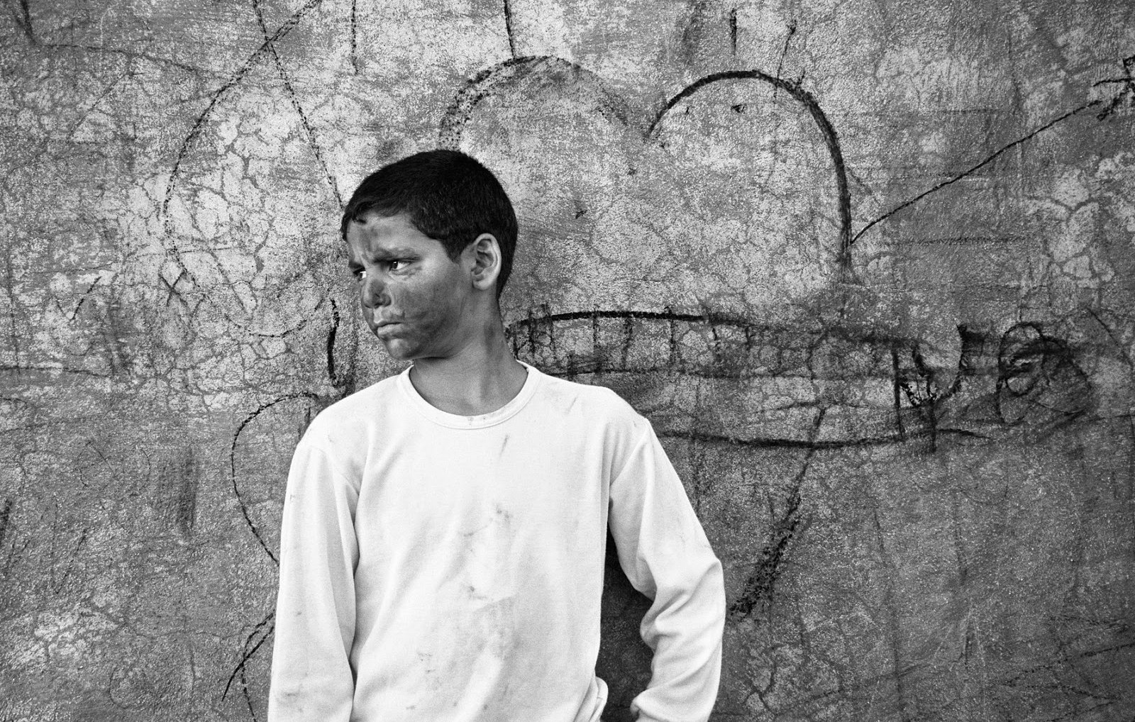 L'image montre un garçon qui se tient contre un mur texturé, où l'on peut voir des éraflures et des graffitis. Il porte un t-shirt blanc légèrement sale et un pantalon beige. Son expression semble pensive ou mélancolique. Le décor derrière lui, avec un dessin de cœur, contraste avec son attitude, créant une atmosphère intéressante. Cela donne une impression de nostalgie ou de réflexion sur son environnement.