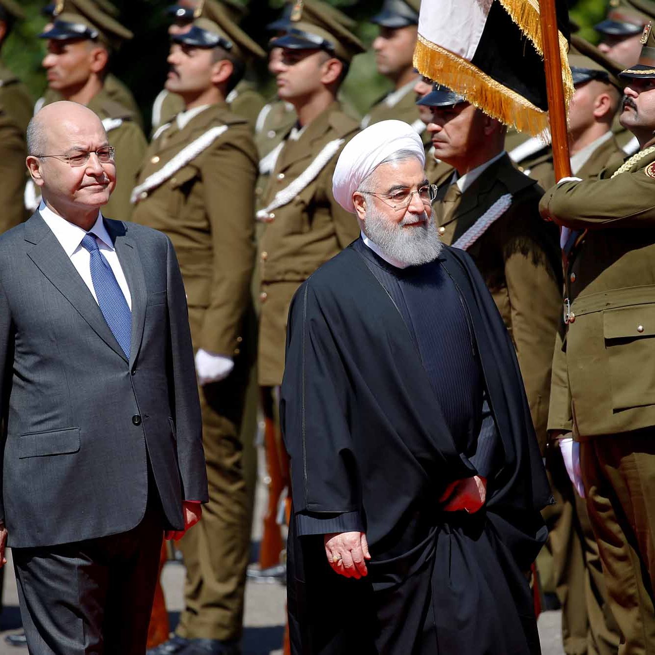 The image shows two men walking in a formal setting, likely during a ceremonial event. One man is dressed in a suit and tie, while the other is wearing a traditional black robe and a white turban. They are surrounded by a group of uniformed soldiers standing at attention, indicating a military or state occasion. The atmosphere appears formal and significant, suggesting a diplomatic meeting or official visit.