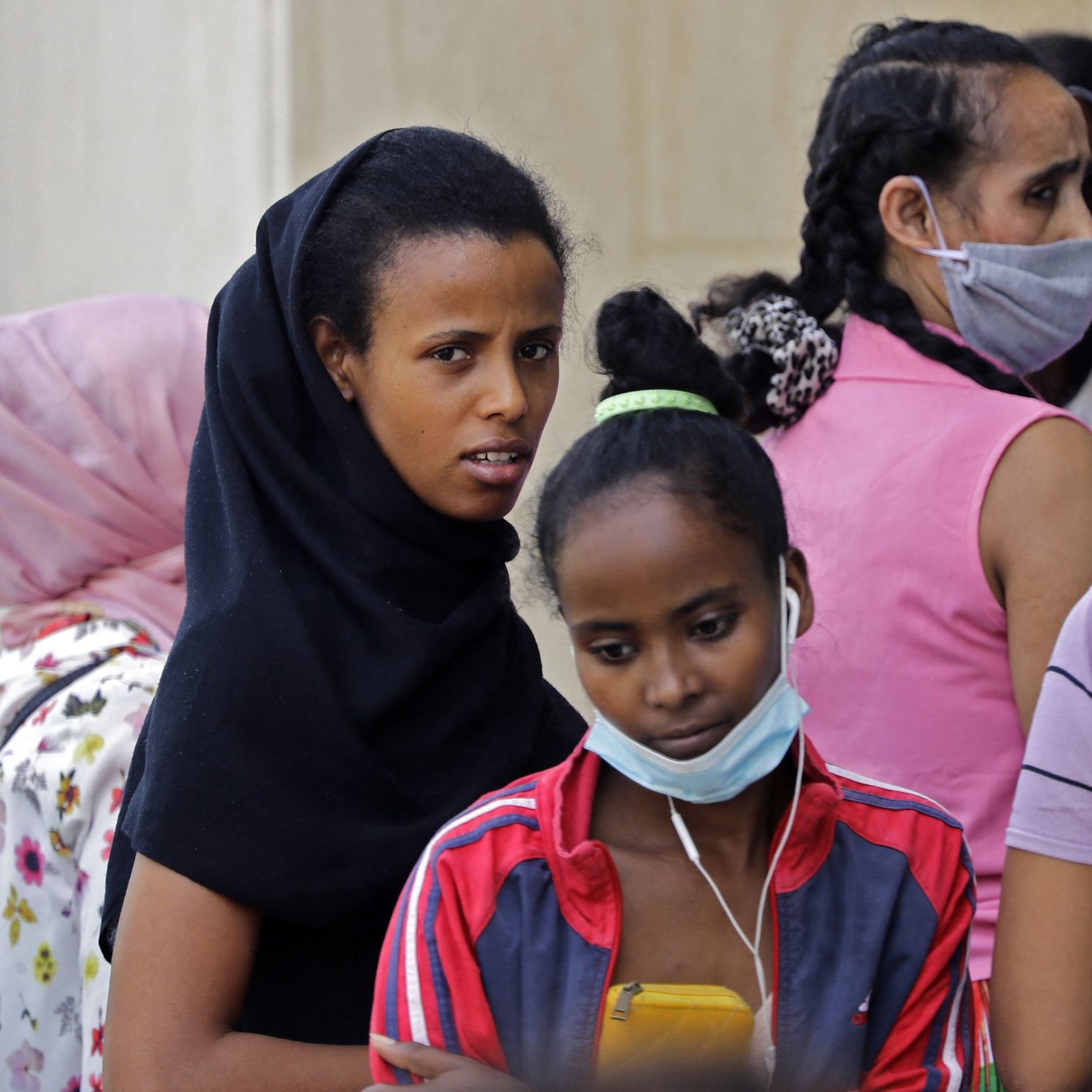 L'image montre un groupe de femmes et de jeunes filles, certaines portant des foulards ou des masques. Les visages expriment divers états d'esprit, allant de la concentration à l'inquiétude. Les vêtements sont colorés, et certaines d'entre elles semblent discuter ou interagir, tandis que d'autres regardent vers l'objectif avec curiosité. L'ambiance générale dénote un moment de rassemblement ou d'attente.